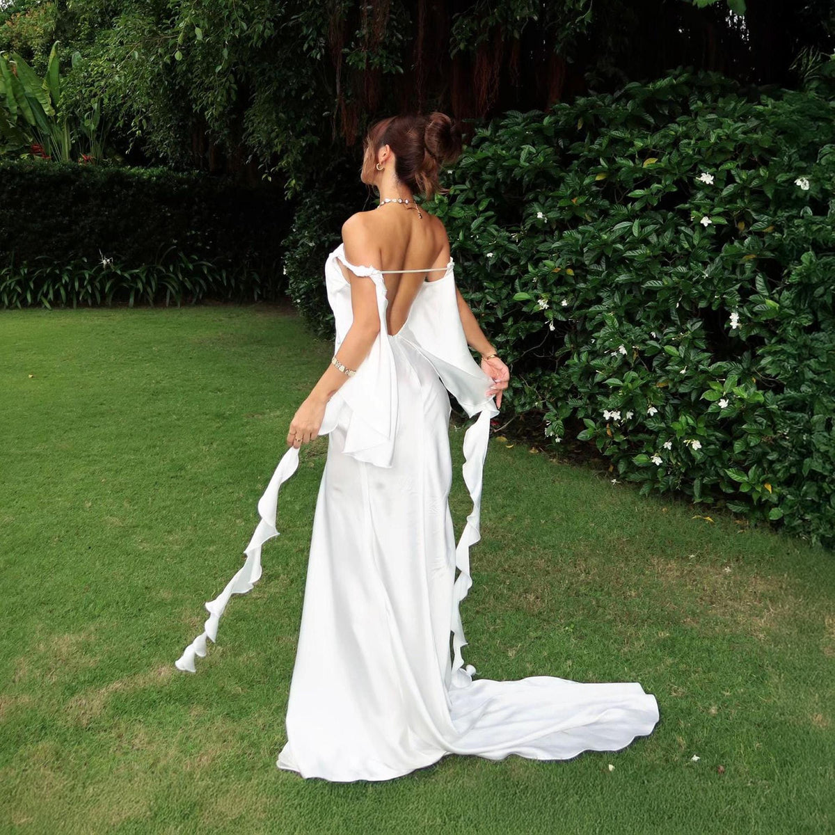 May include: A floor-length white satin gown featuring an open back, off-the-shoulder sleeves, and long, cascading ruffled fabric panels that extend from the shoulders down to the floor, creating a flowing silhouette with a train.