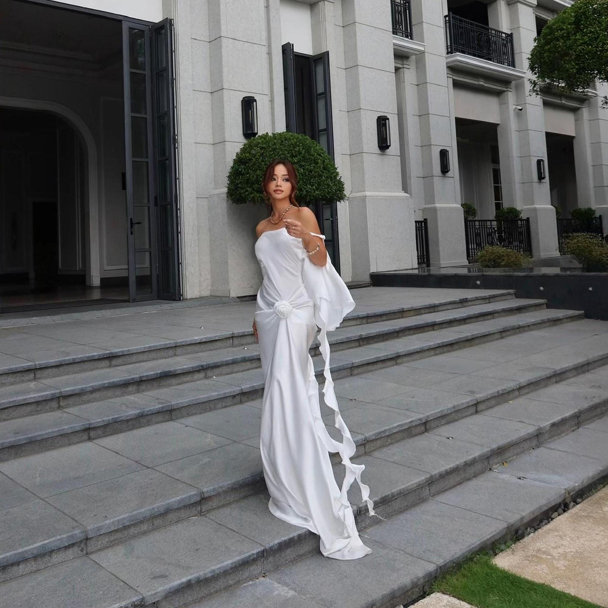 May include: A floor-length white off-the-shoulder dress featuring a draped bodice with a central fabric rose detail, a fitted silhouette, and a long, flowing sleeve extension with a ruffled edge that cascades down to the floor.