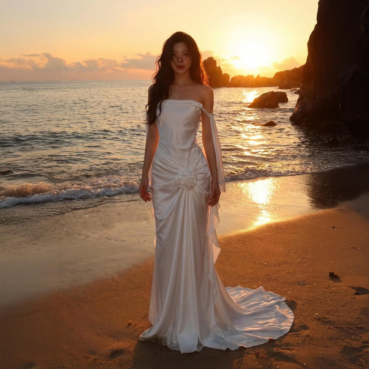 May include: A floor-length white strapless gown featuring a smooth satin finish, a gathered waist detail with a central fabric rosette, and long, flowing off-the-shoulder sleeves that drape down the sides, ending in a slight train on the sand.