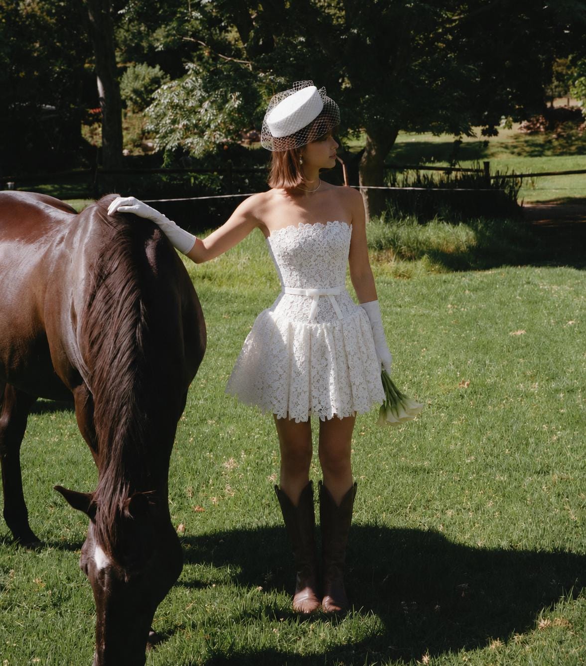 May include: A white strapless lace mini dress with a thin waist belt, paired with white elbow-length gloves, a white pillbox hat with a birdcage veil, and dark brown leather cowboy boots. The ensemble includes a small bouquet of white calla lilies.