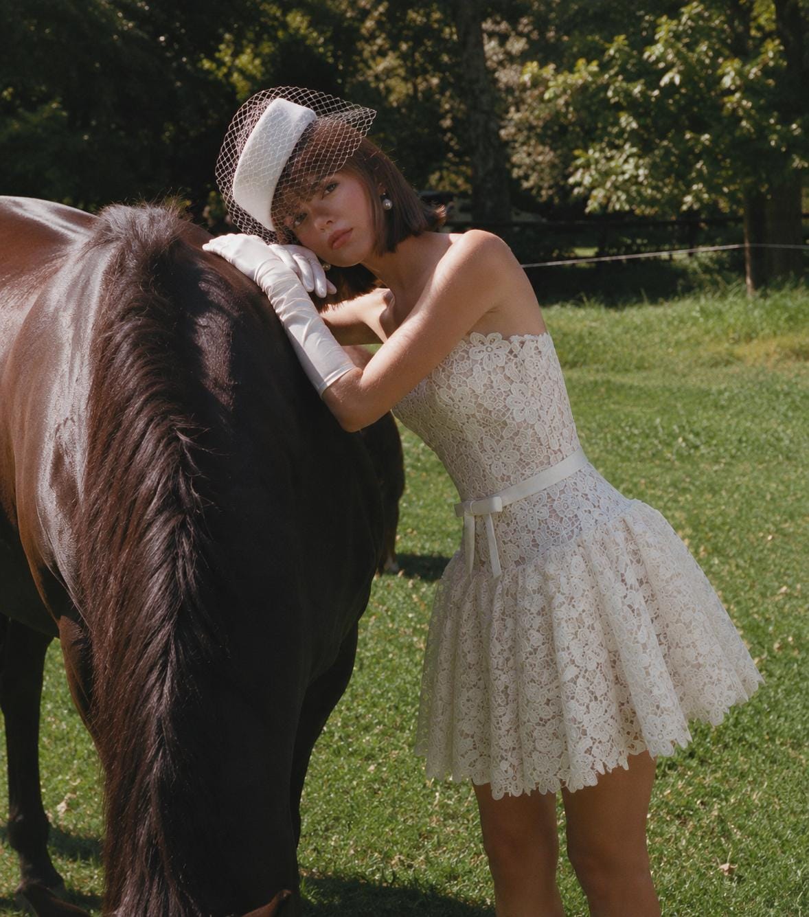 May include: A strapless white lace mini dress featuring a fitted bodice with a thin waist ribbon and a flared skirt. The ensemble includes white elbow-length gloves and a white pillbox hat adorned with a delicate netting veil.