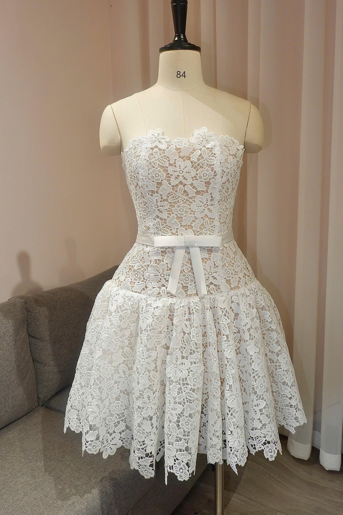 May include: A strapless white lace mini dress featuring a floral pattern over a nude-colored lining. The dress has a fitted bodice and a flared skirt, accented at the waist with a smooth white satin ribbon tied into a bow at the center front.