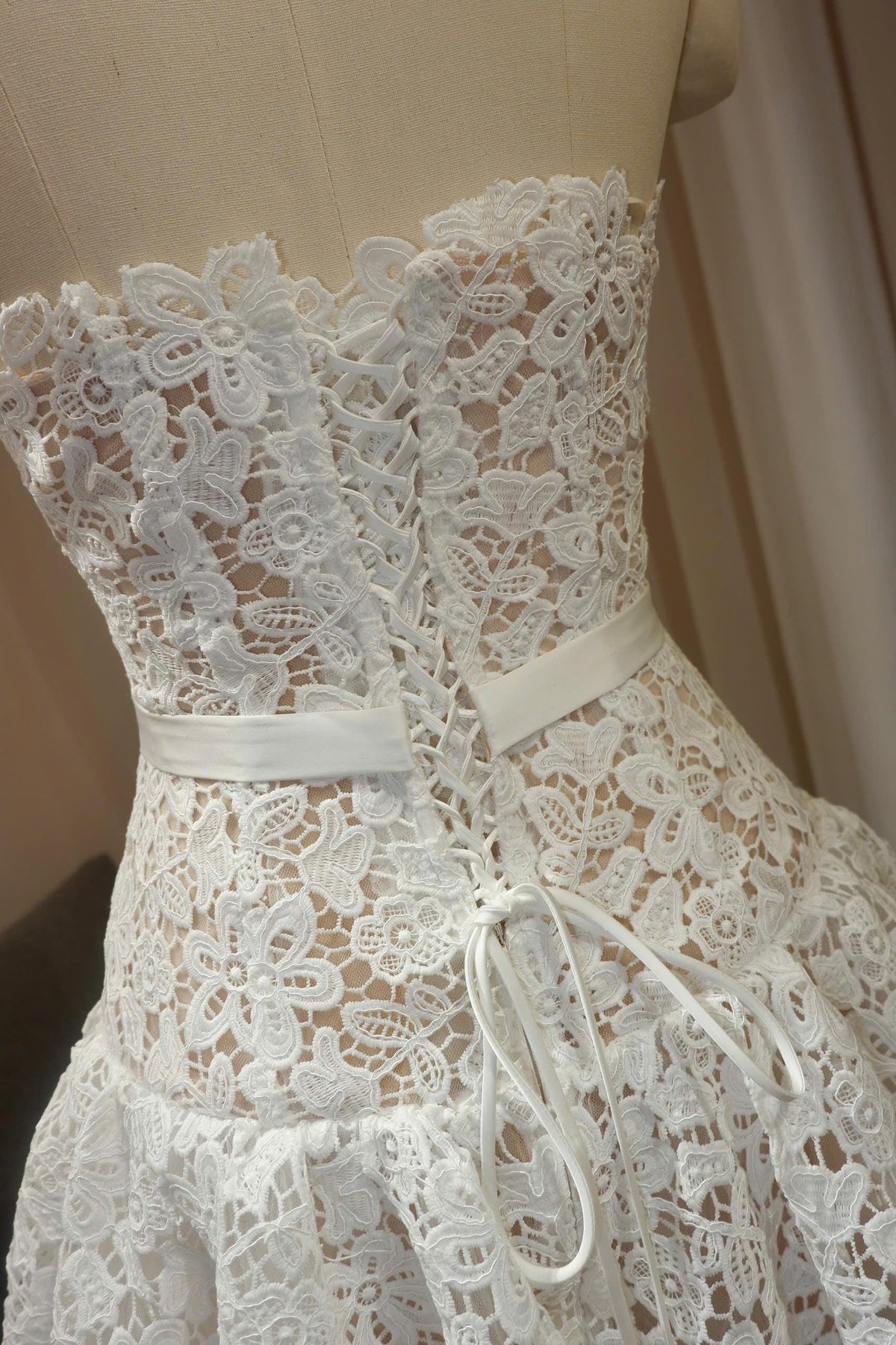 May include: A strapless white lace dress featuring a floral pattern and a corset-style back with a lace-up ribbon closure. The bodice includes a smooth white satin waistband that sits above a flared skirt, showcasing intricate embroidery.