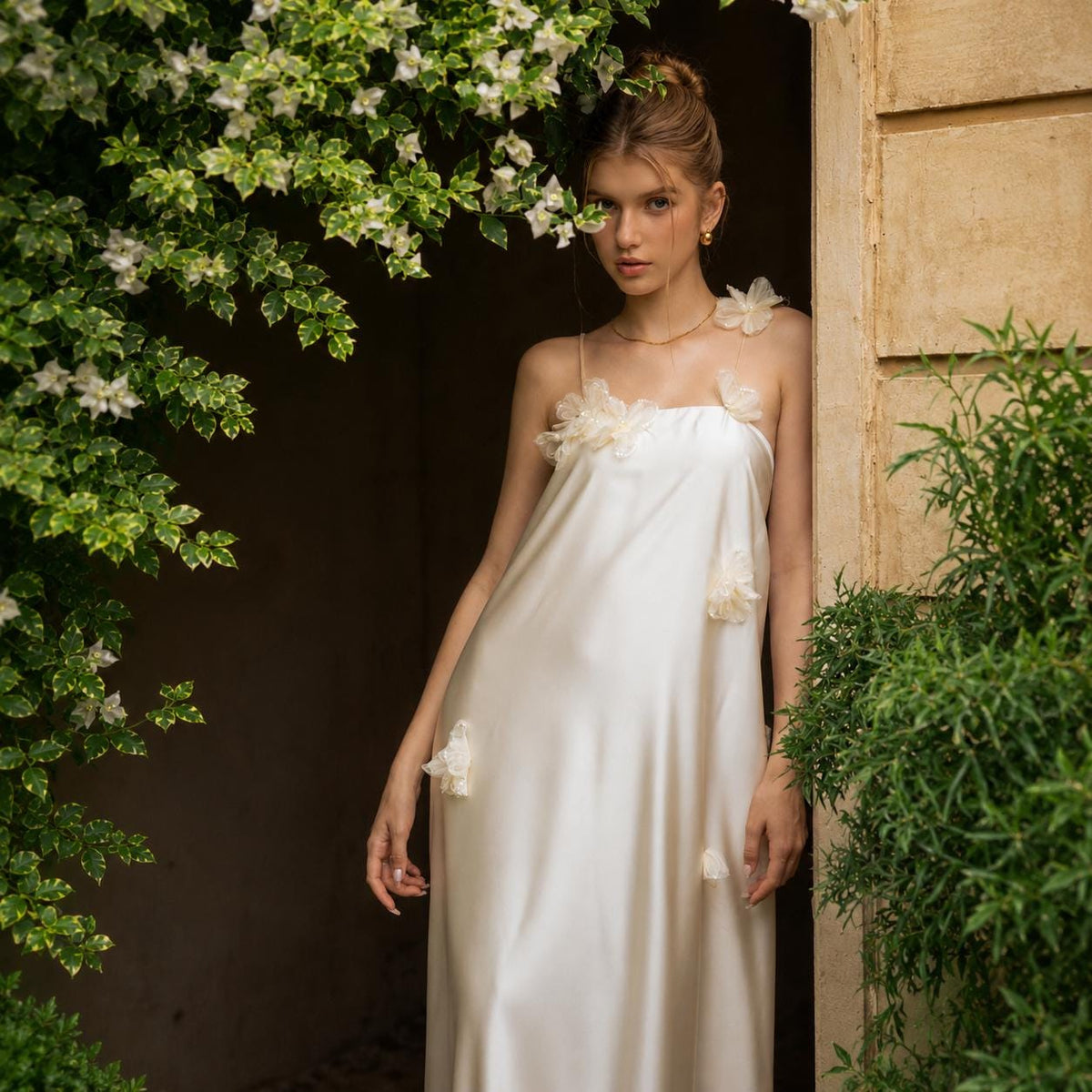 May include: A floor-length, cream-colored satin slip dress featuring thin spaghetti straps and delicate, three-dimensional fabric flower appliqués scattered across the bodice and skirt. The garment has a straight neckline and a fluid silhouette.