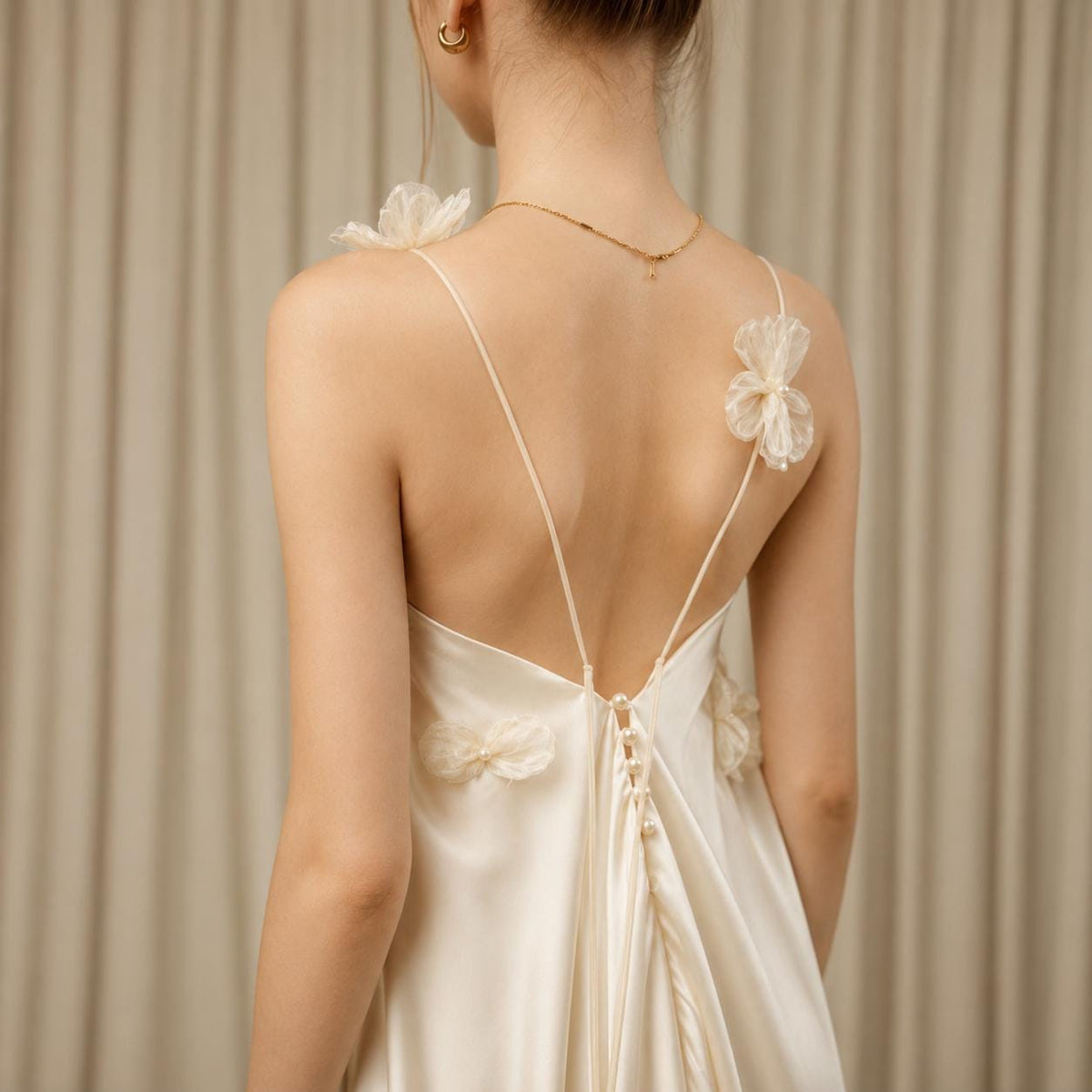 May include: A cream-colored satin slip dress featuring a low back with thin spaghetti straps. The dress is adorned with delicate, sheer fabric floral appliqués and a vertical row of small, round white pearl buttons along the center back seam.