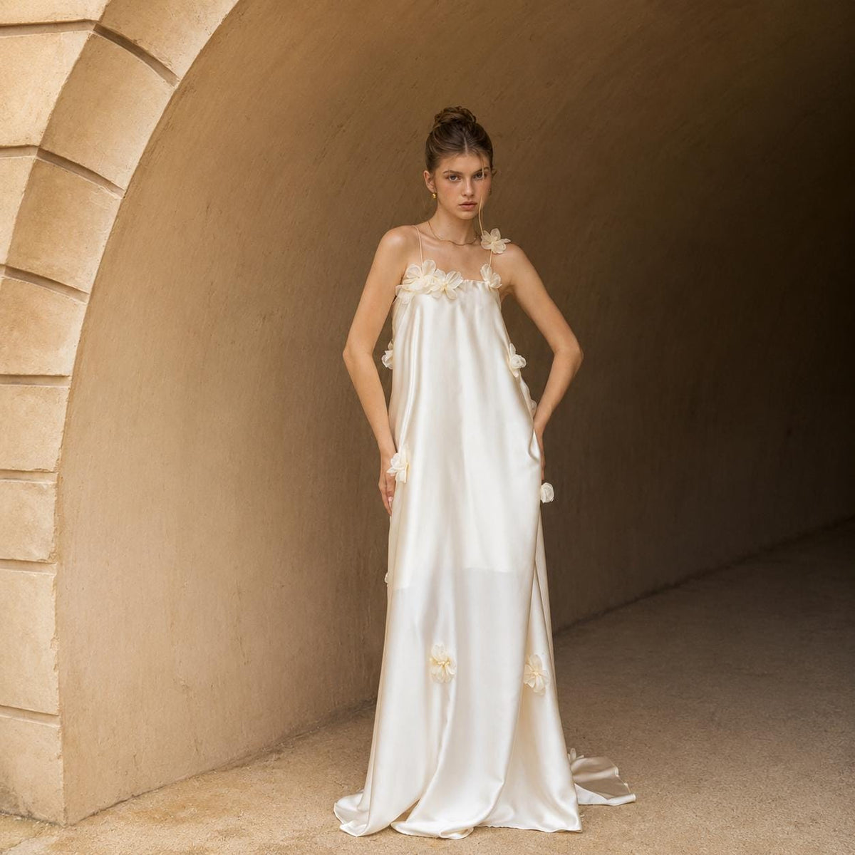 May include: A floor-length cream-colored satin dress featuring thin spaghetti straps and a straight neckline. The garment is adorned with scattered three-dimensional fabric flower appliqués in a matching cream tone, creating a textured surface.