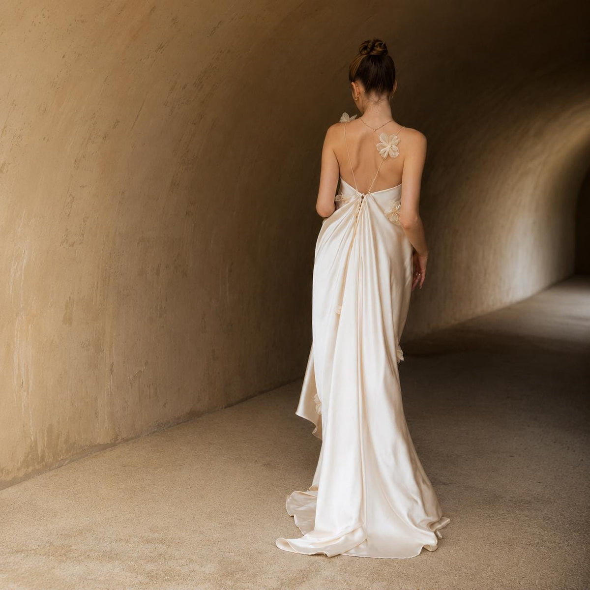 May include: A floor-length cream-colored satin gown featuring a backless design with thin straps, a row of small buttons along the center back, and delicate three-dimensional floral appliqués attached to the shoulder and side of the skirt.