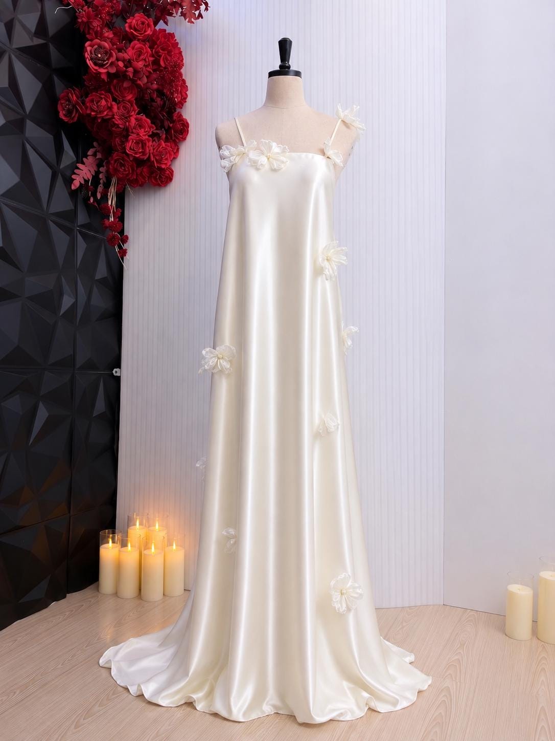 May include: A floor-length cream-colored satin dress featuring thin spaghetti straps and a straight neckline. The garment is adorned with scattered three-dimensional fabric flower appliqués in a matching cream tone across the bodice and skirt.