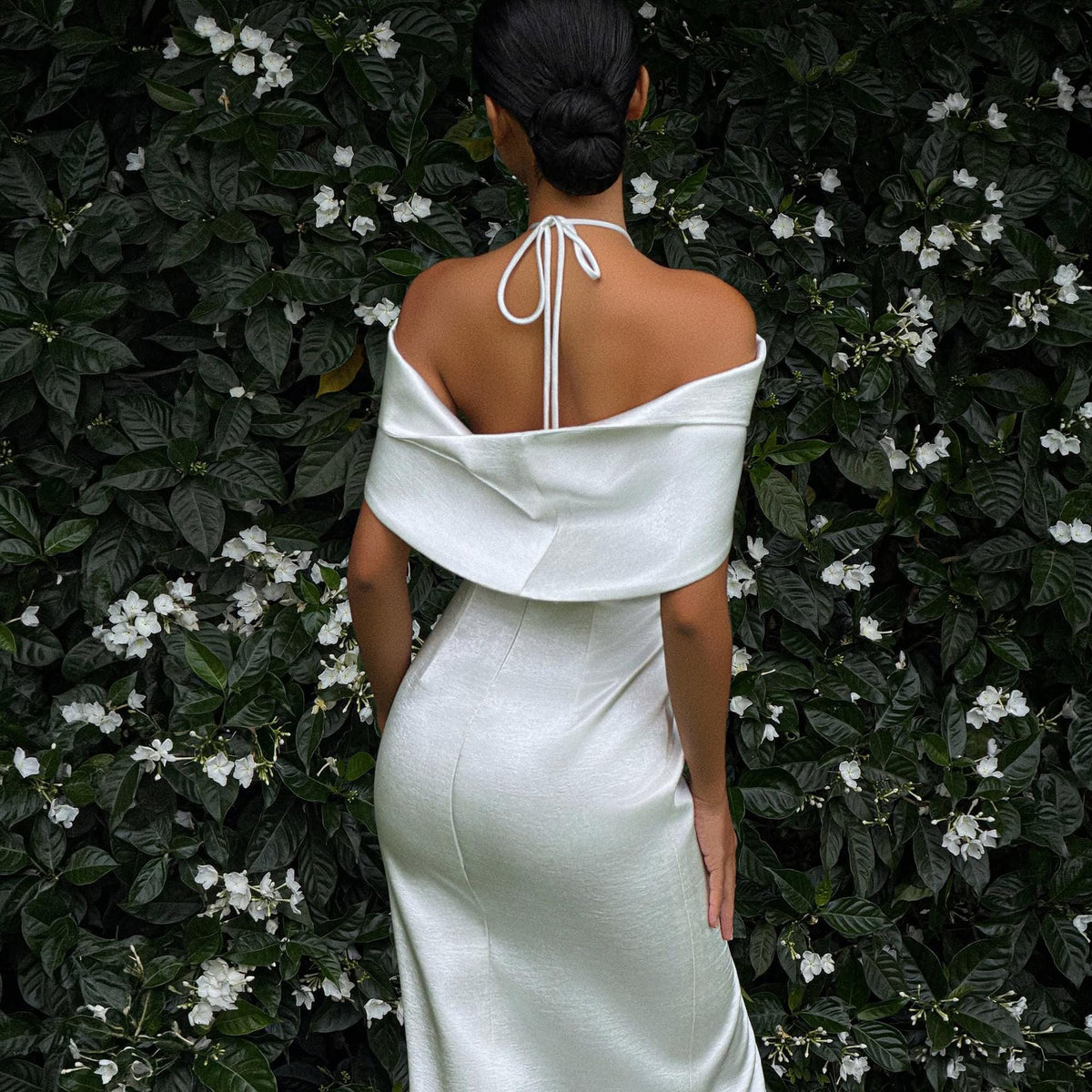 May include: A floor-length white dress featuring an off-the-shoulder neckline with a draped fold-over design. The garment includes thin white halter-style straps that tie at the back of the neck, creating a smooth, fitted silhouette.