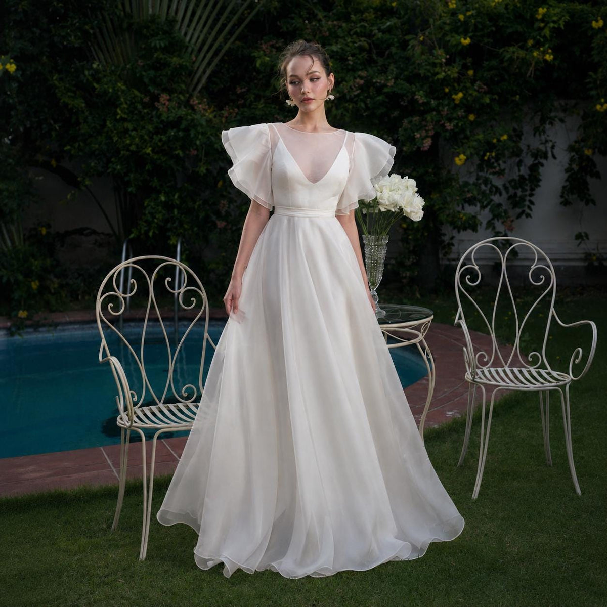 May include: A floor-length white gown featuring a sheer illusion neckline, a fitted bodice, and voluminous ruffled flutter sleeves. The skirt is made of a flowing, layered fabric that extends to the ground, creating a soft and elegant silhouette.