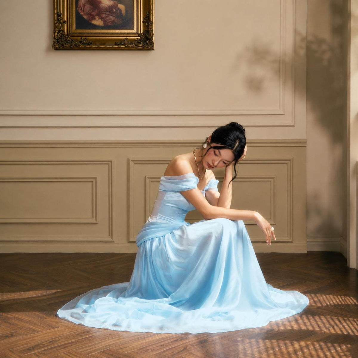 May include: A floor-length light blue gown featuring an off-the-shoulder neckline with draped sleeves and a fitted bodice. The skirt is made of a flowing, sheer fabric that pools on the floor, creating a soft silhouette with layered material.
