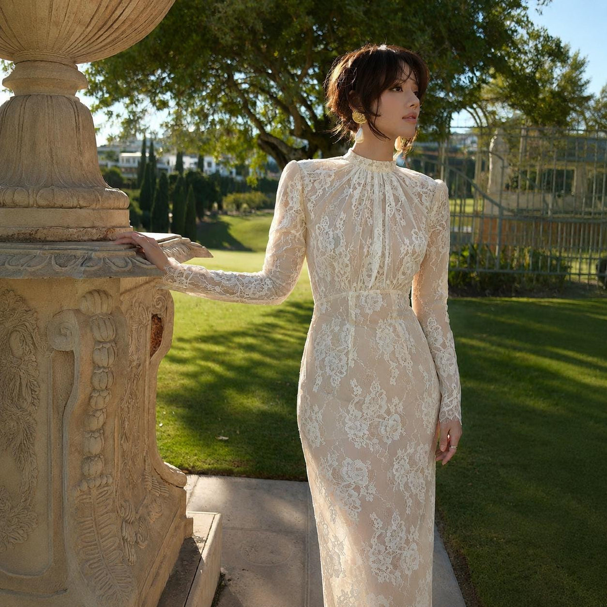 May include: A floor-length dress featuring a high neckline and long sleeves, crafted from white floral lace layered over a nude-colored fabric. The garment has a fitted bodice and a straight skirt silhouette, showcasing intricate lace patterns throughout.