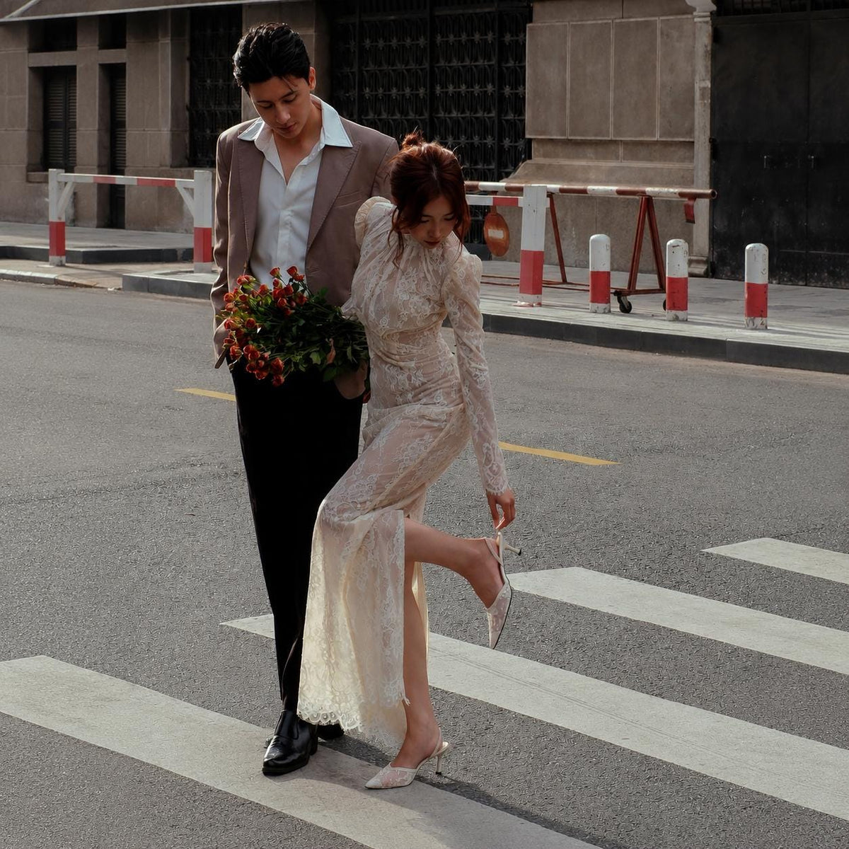 May include: A beige blazer, white button-down shirt, and black trousers paired with a long-sleeved, floor-length cream lace dress featuring a high neckline and a side slit. A bouquet of red roses is held alongside the cream lace slingback heels.