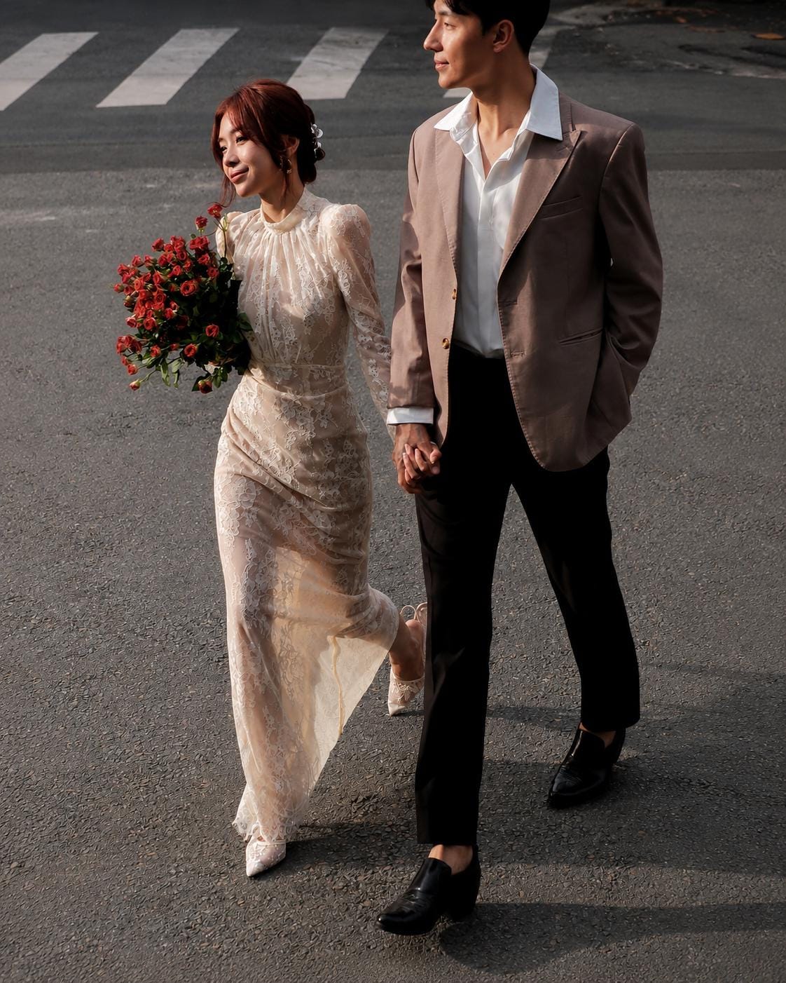 May include: A long-sleeved cream lace dress with a high neckline and a floor-length skirt, paired with a tan blazer, a white collared shirt, and black trousers. A bouquet of red roses is held in one hand while walking on an asphalt surface.