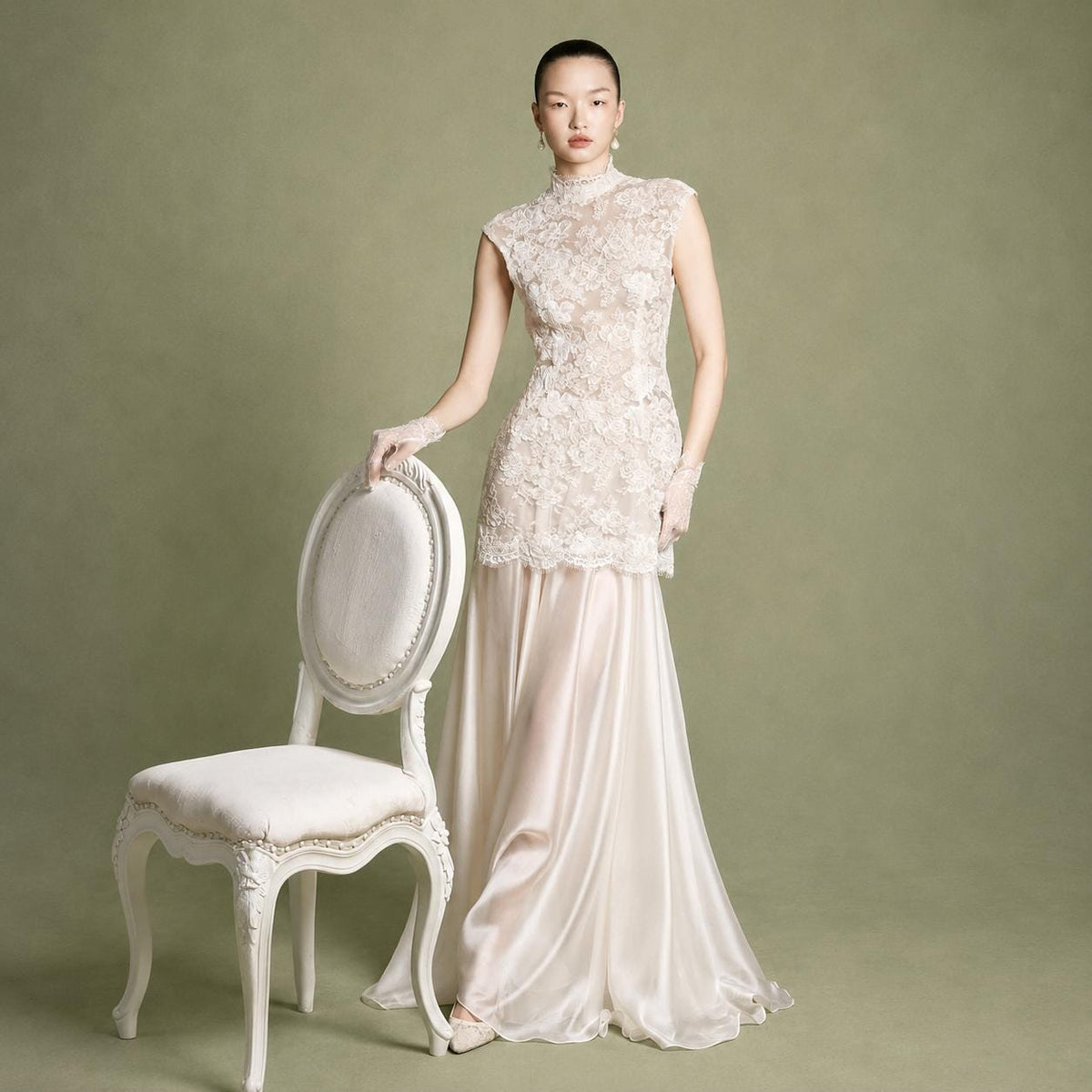 May include: A floor-length gown featuring a sleeveless, high-neck bodice covered in intricate cream-colored floral lace overlay. The skirt is made of a smooth, flowing satin fabric in a matching cream tone, creating a layered appearance.