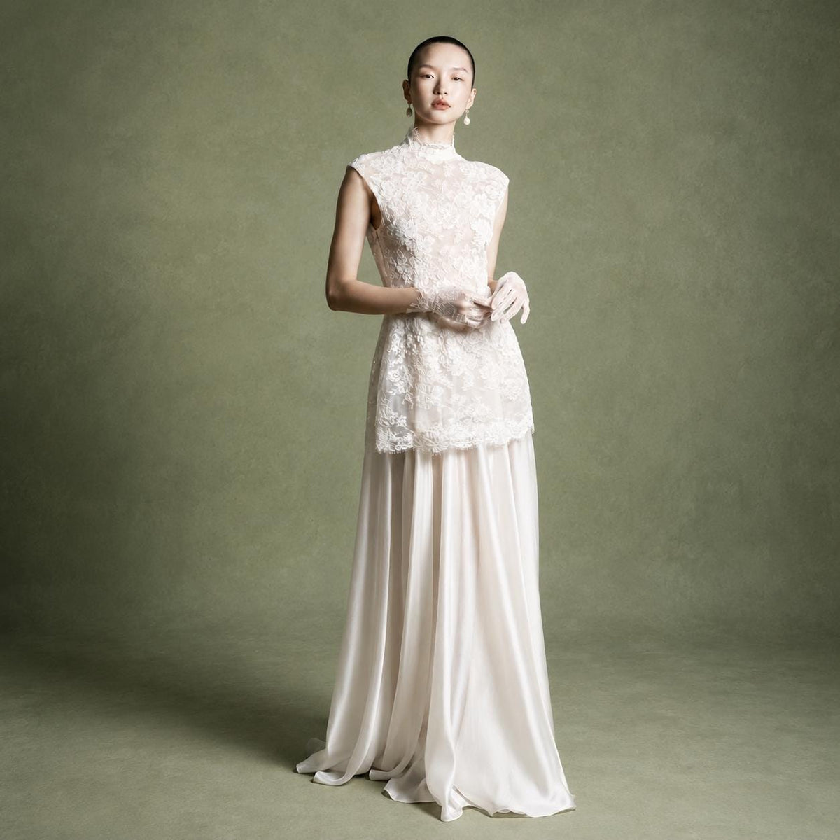 May include: A floor-length gown featuring a sleeveless, high-neck bodice crafted from intricate white lace with a scalloped hem. The skirt is made of a smooth, flowing off-white satin fabric that drapes to the floor. Matching lace gloves are worn.