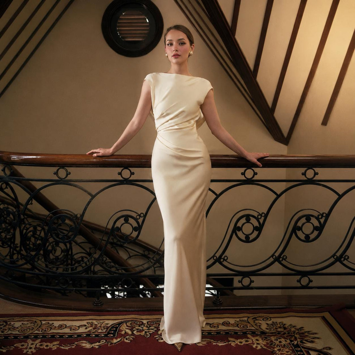 May include: A floor-length, cream-colored sleeveless gown featuring a boat neckline and intricate draping at the waist. The garment is crafted from a smooth, satin-like fabric that flows down to the floor, creating a sleek and form-fitting silhouette.