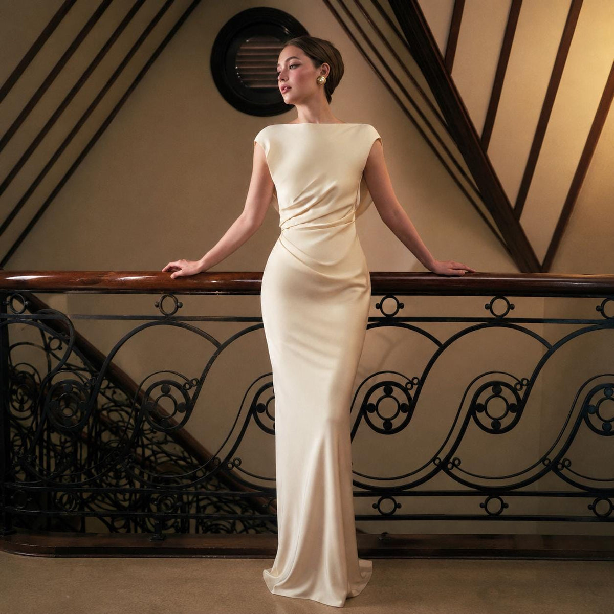 May include: A floor-length, cream-colored gown featuring a boat neckline, cap sleeves, and subtle side-waist ruching. The dress is crafted from a smooth, satin-like fabric that drapes down to the floor in a straight, form-fitting silhouette.