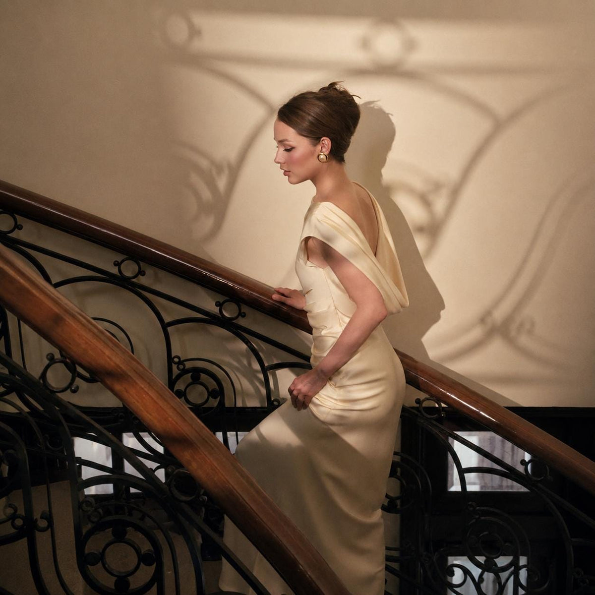 May include: A floor-length cream-colored gown featuring a draped neckline, short sleeves, and a fitted silhouette. The dress is made of a smooth, light-reflecting fabric that falls to the floor, showcasing a clean, simple design with no patterns.