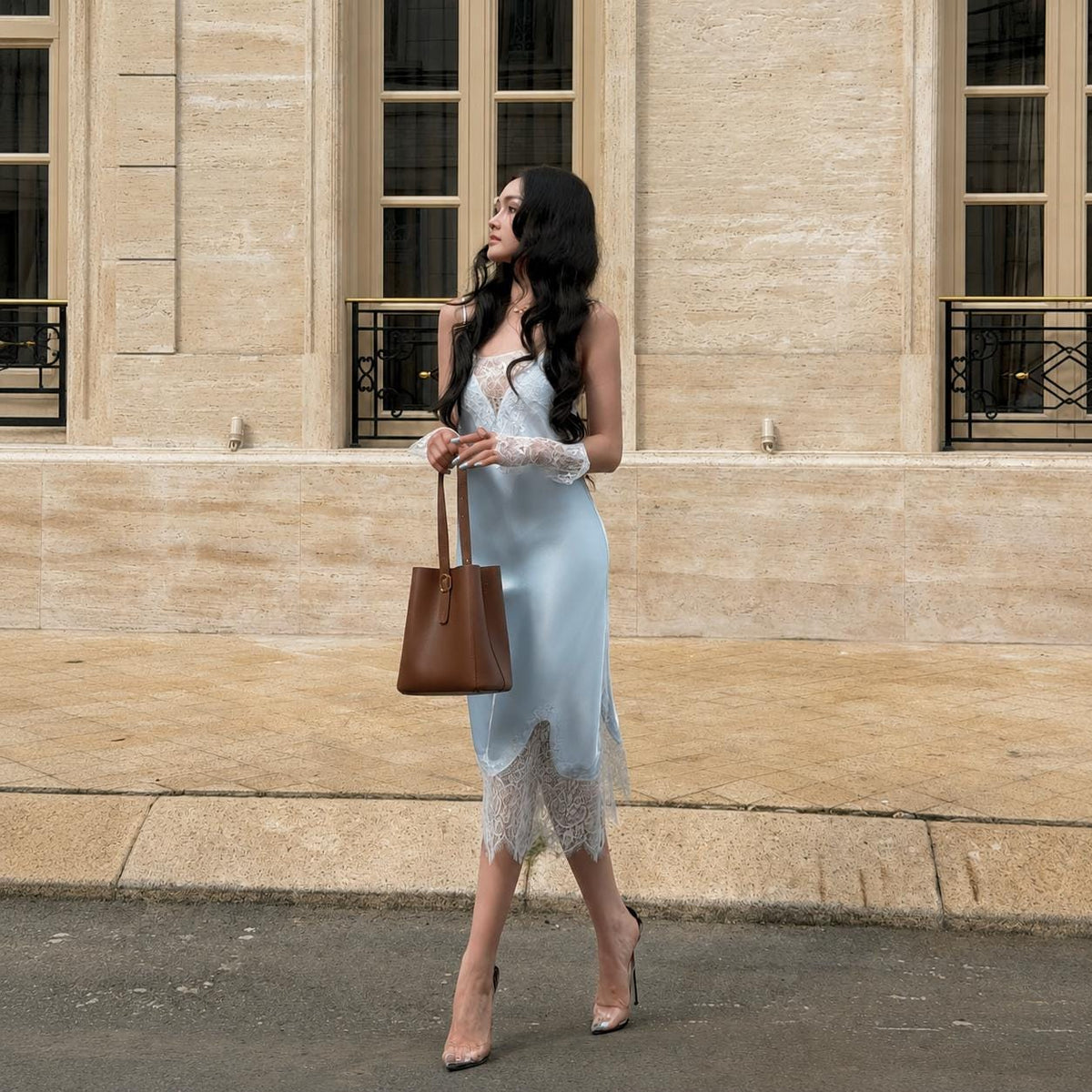 May include: A light blue satin midi dress featuring a white lace bodice overlay and a matching white lace hemline. The dress is paired with a brown leather bucket bag and clear high-heeled shoes, creating a coordinated ensemble with delicate lace details.