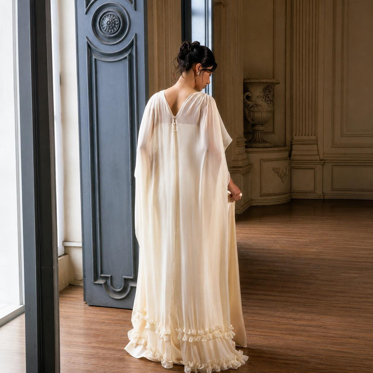 May include: A floor-length cream-colored gown featuring a sheer, flowing cape-style overlay that drapes from the shoulders. The dress has a V-neckline at the back, a central vertical seam, and a ruffled hemline that adds texture to the bottom edge.