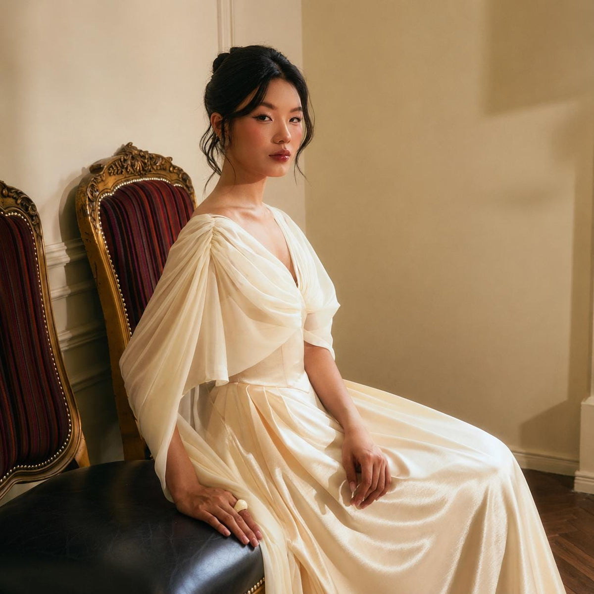May include: A floor-length cream-colored gown featuring a V-neckline, gathered fabric at the shoulders, and flowing, draped sleeves. The dress is constructed from a lightweight, semi-sheer material that falls into a full, smooth skirt.