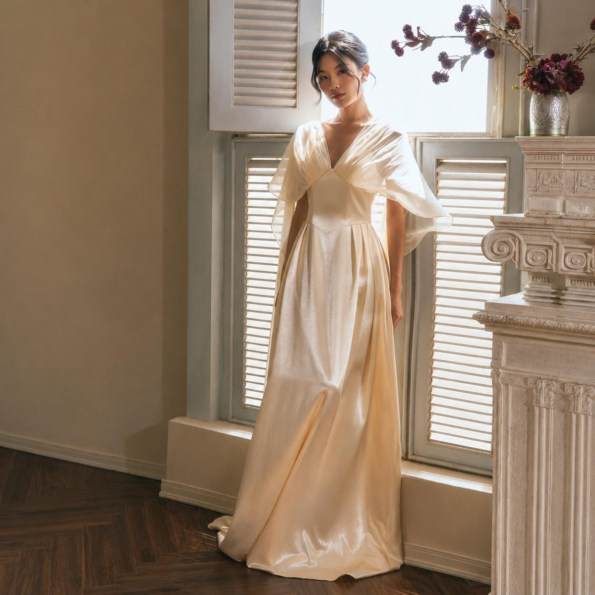 May include: A floor-length champagne-colored satin gown featuring a V-neckline, a fitted bodice, and sheer, flowing cape-style sleeves that drape over the shoulders. The skirt is pleated at the waist, creating a full, elegant silhouette that falls to the floor.