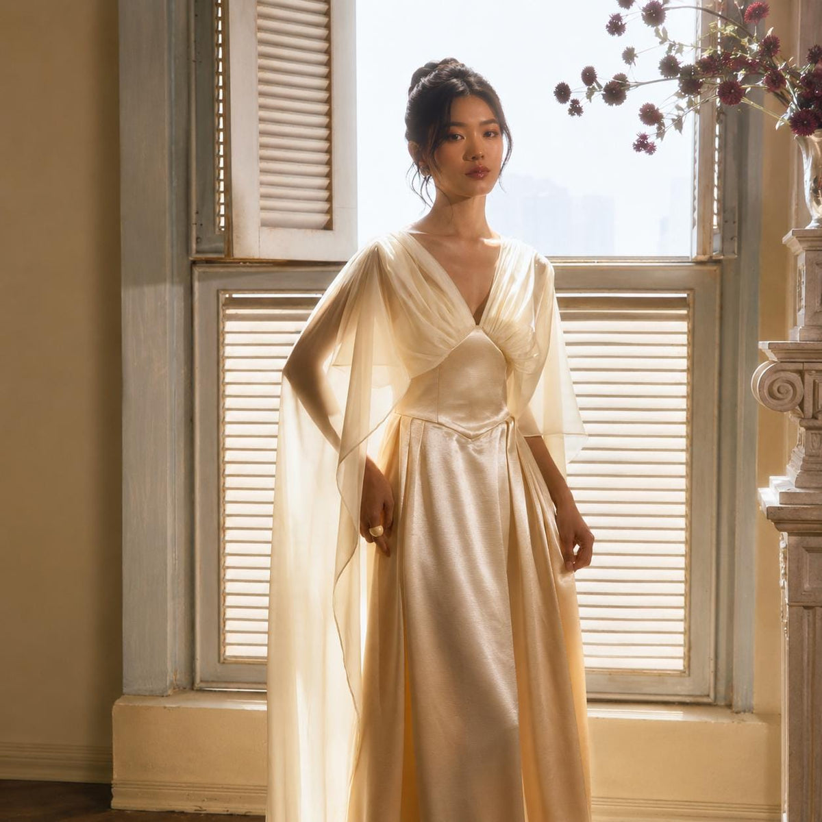 May include: A floor-length champagne-colored satin gown featuring a deep V-neckline, a fitted bodice with a defined waist, and flowing, sheer cape-style sleeves that drape from the shoulders. The fabric has a smooth, lustrous finish.
