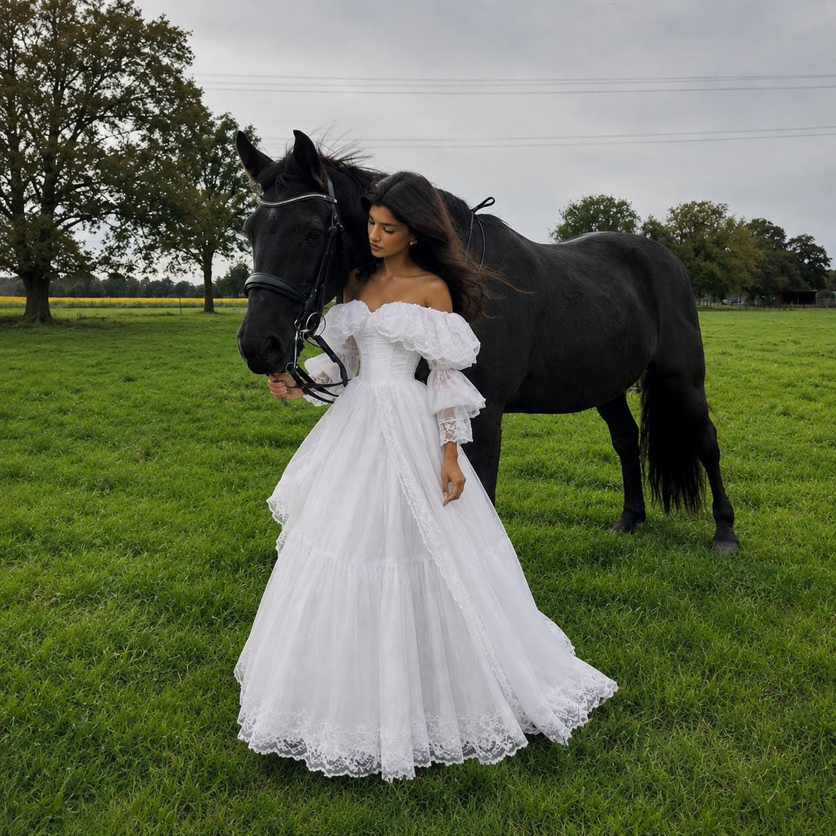 May include: A white off-the-shoulder gown featuring voluminous puff sleeves, a fitted bodice, and a floor-length skirt with tiered layers and intricate lace trim along the hem, standing beside a large black horse in a green grassy field.