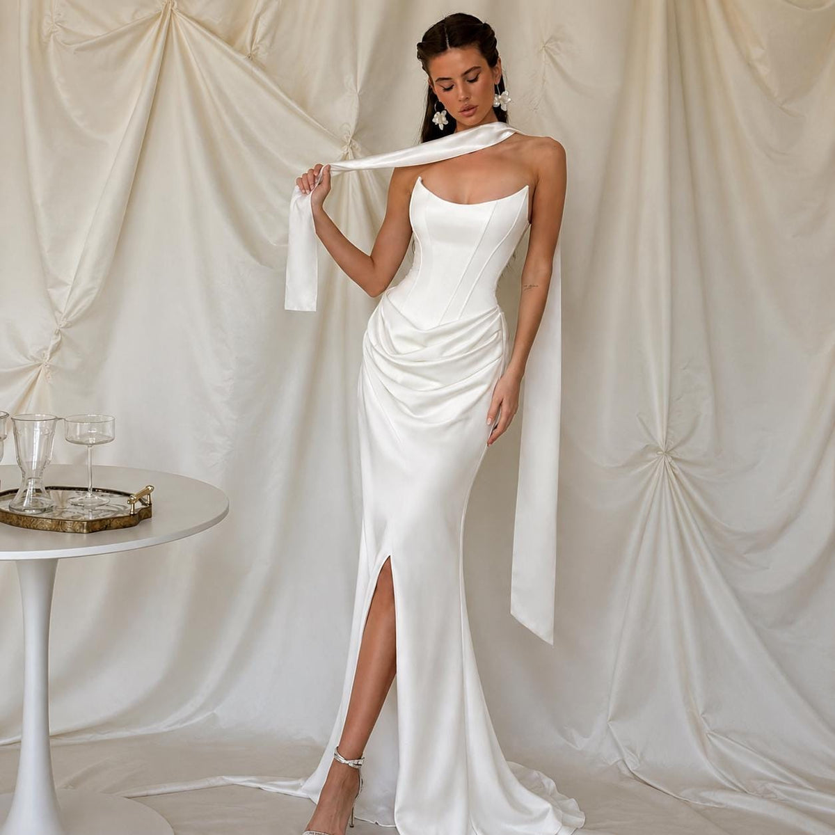 May include: A floor-length white satin gown featuring a structured corset-style bodice with a scoop neckline, a draped skirt with a high front slit, and a long, flowing scarf-like attachment draped over the shoulders and down the sides.