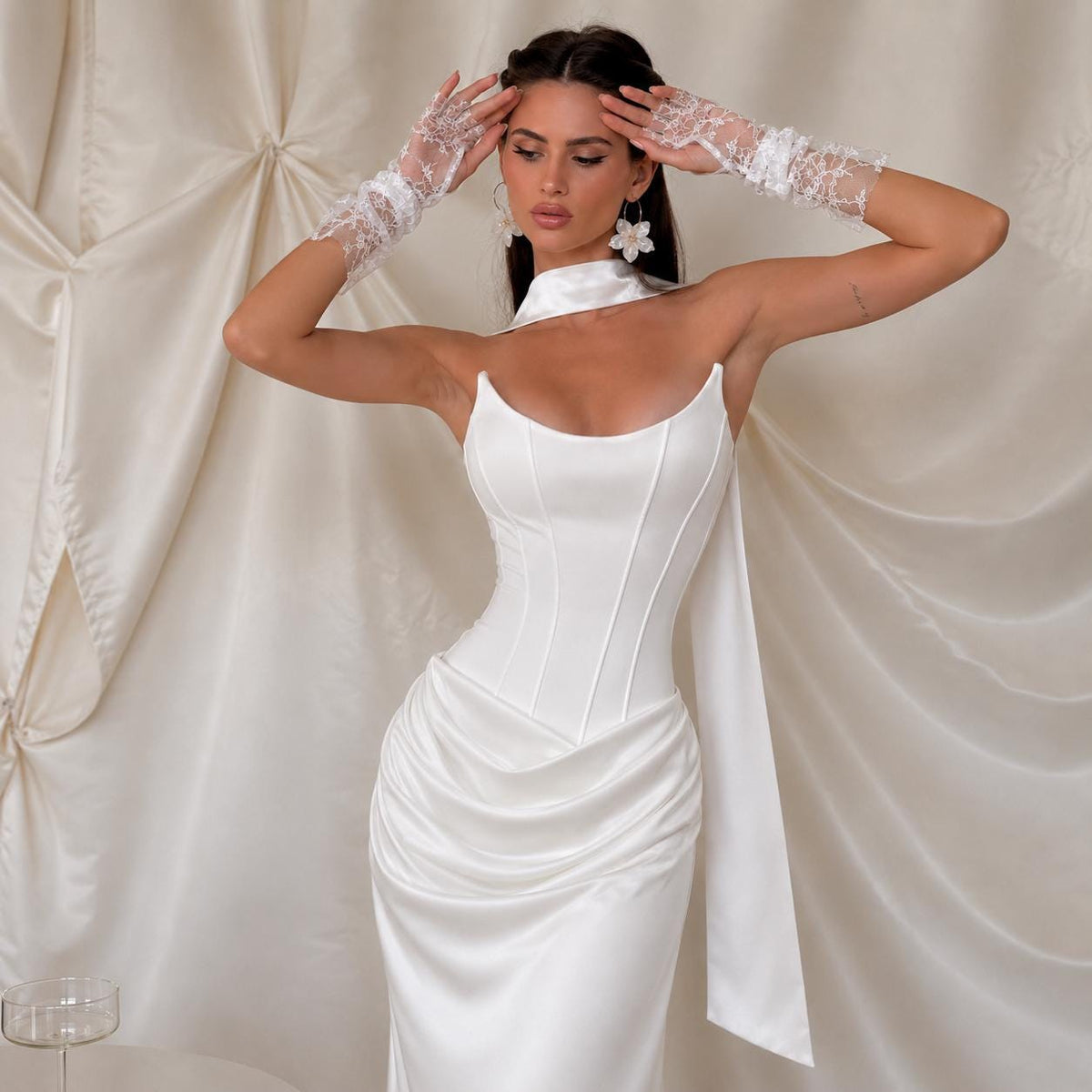 May include: A white satin corset dress featuring a structured bodice with vertical seams, a draped skirt, and a high neck collar. The ensemble includes matching white lace fingerless gloves that extend to the mid-forearm and floral earrings.