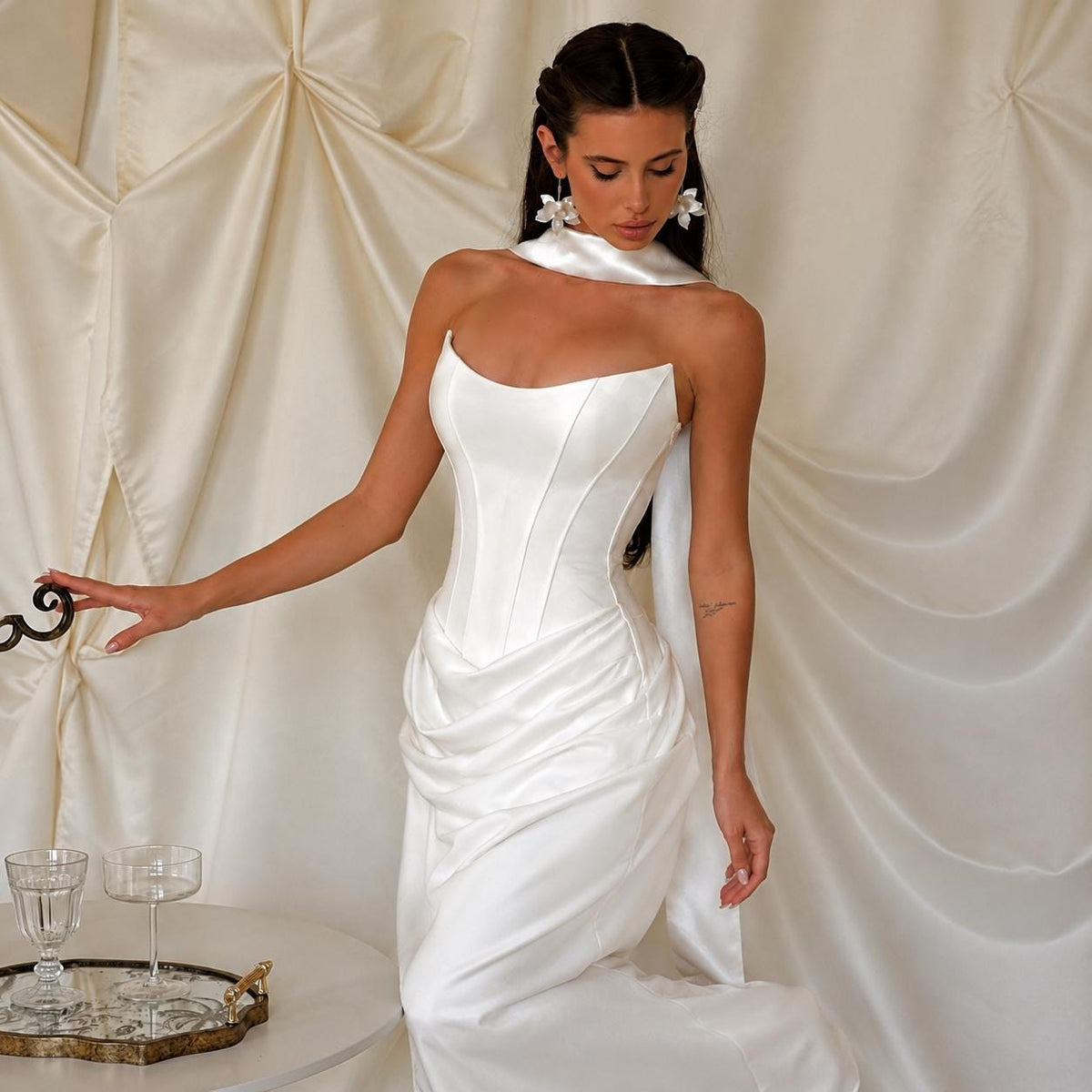 May include: A floor-length white satin gown featuring a structured corset-style bodice with vertical seams, a sweetheart neckline, and a draped, gathered skirt. The dress includes a matching white satin neck scarf and floral-shaped earrings.