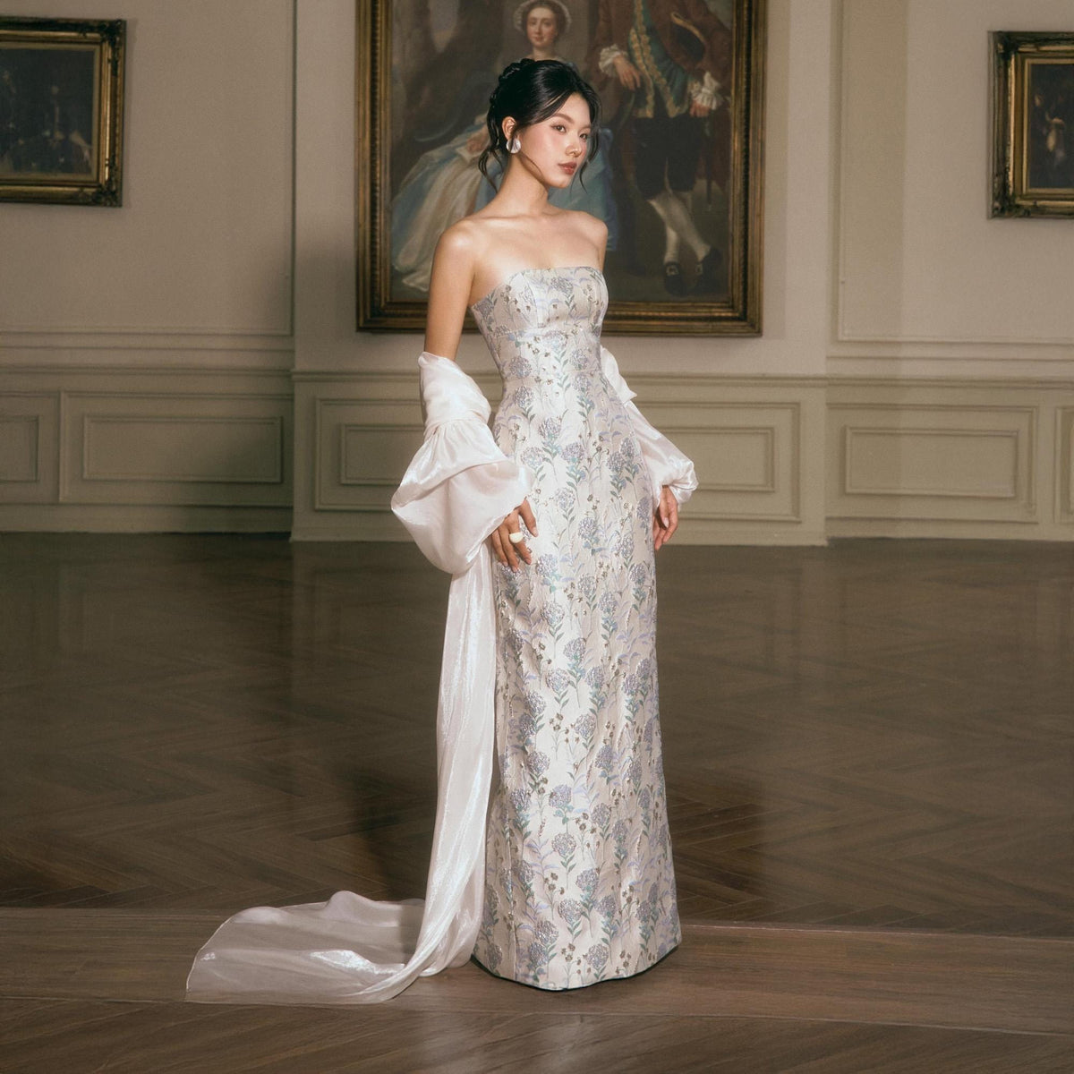 May include: A strapless floor-length gown featuring a light-colored fabric with a repeating blue and silver floral pattern. The dress is paired with a long, flowing white satin shawl that drapes from the arms down to the floor in a train.