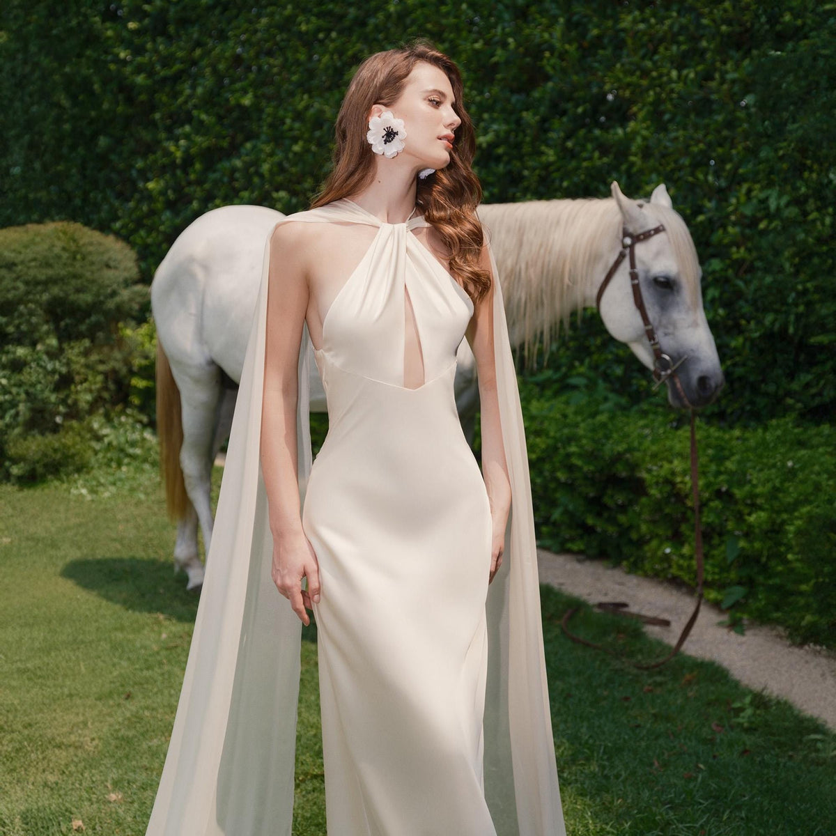 May include: A woman wearing a white halter-neck gown with a long cape, standing in a garden with a white horse in the background.