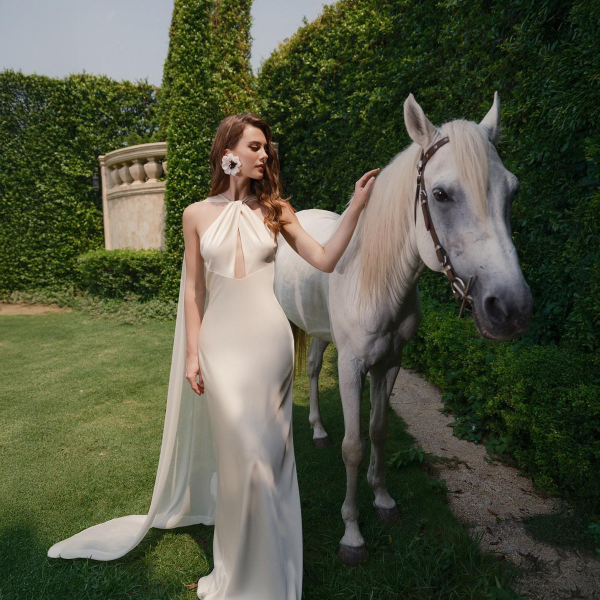 May include: A woman in a white halter-neck gown with a long train stands next to a white horse in a grassy garden setting.