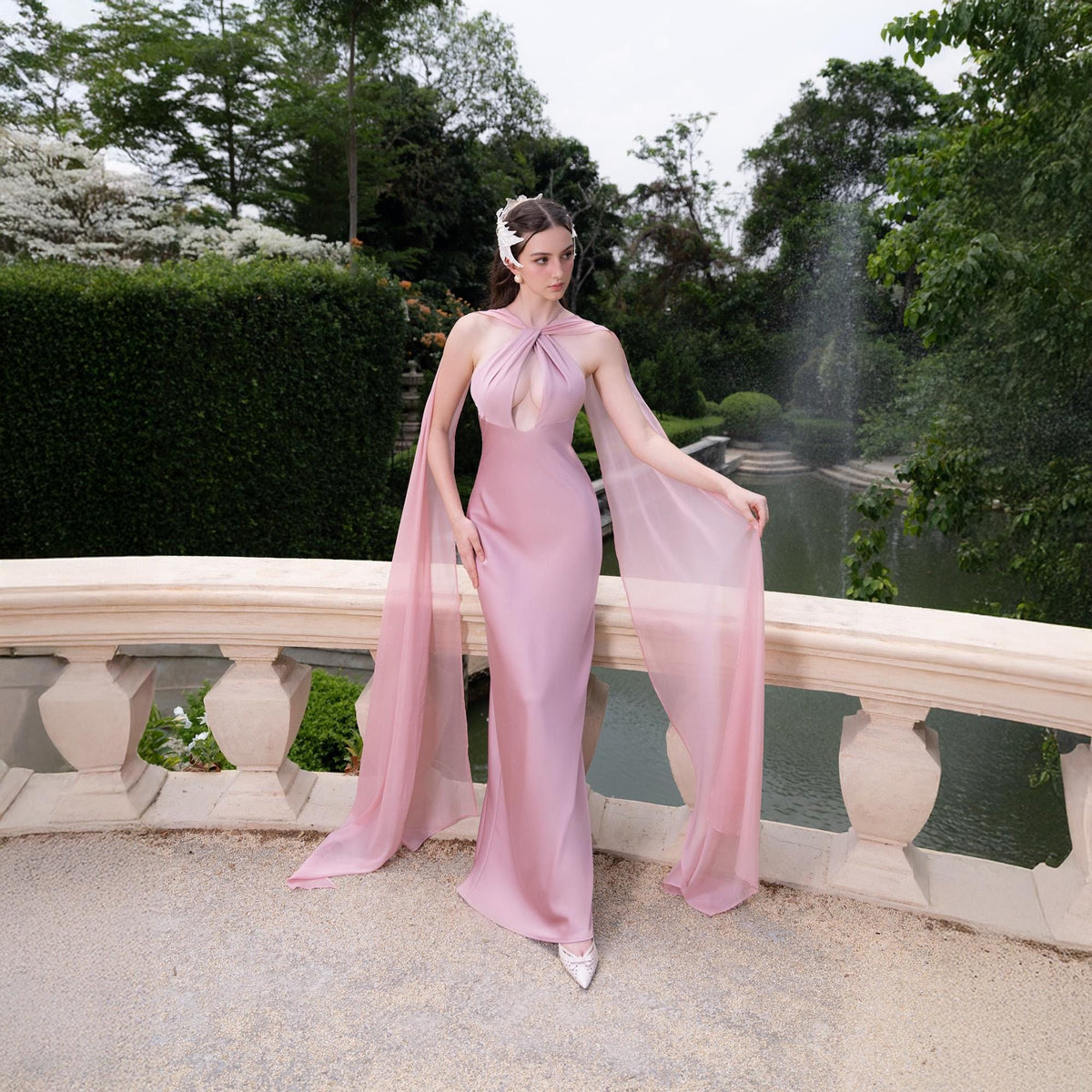 May include: A woman wearing a pink satin gown with a sheer pink cape. The gown has a plunging neckline and a fitted silhouette. The woman is standing on a stone balcony with a garden in the background.
