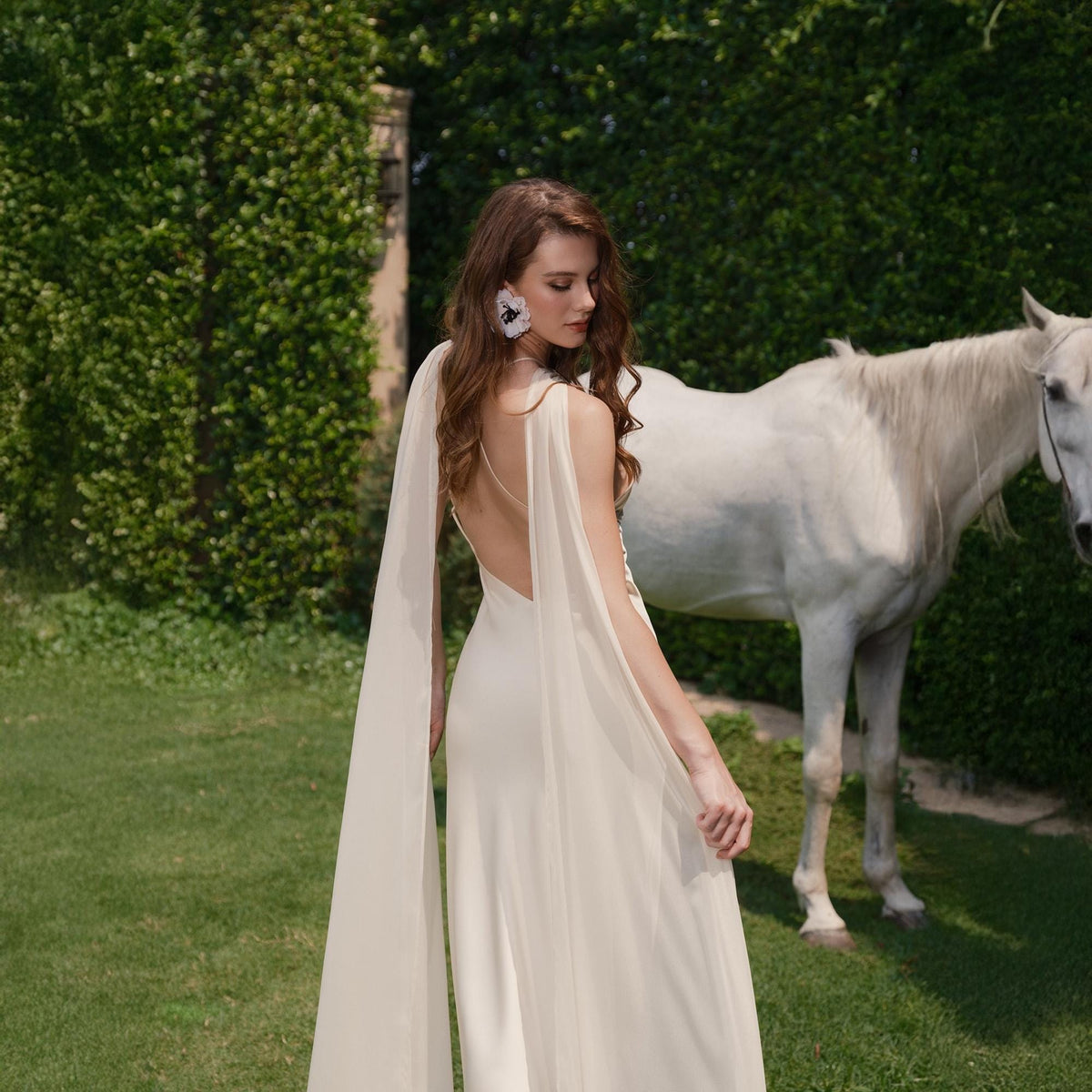 May include: A woman wearing a white sleeveless gown with a sheer cape, standing in a grassy field with a white horse in the background.