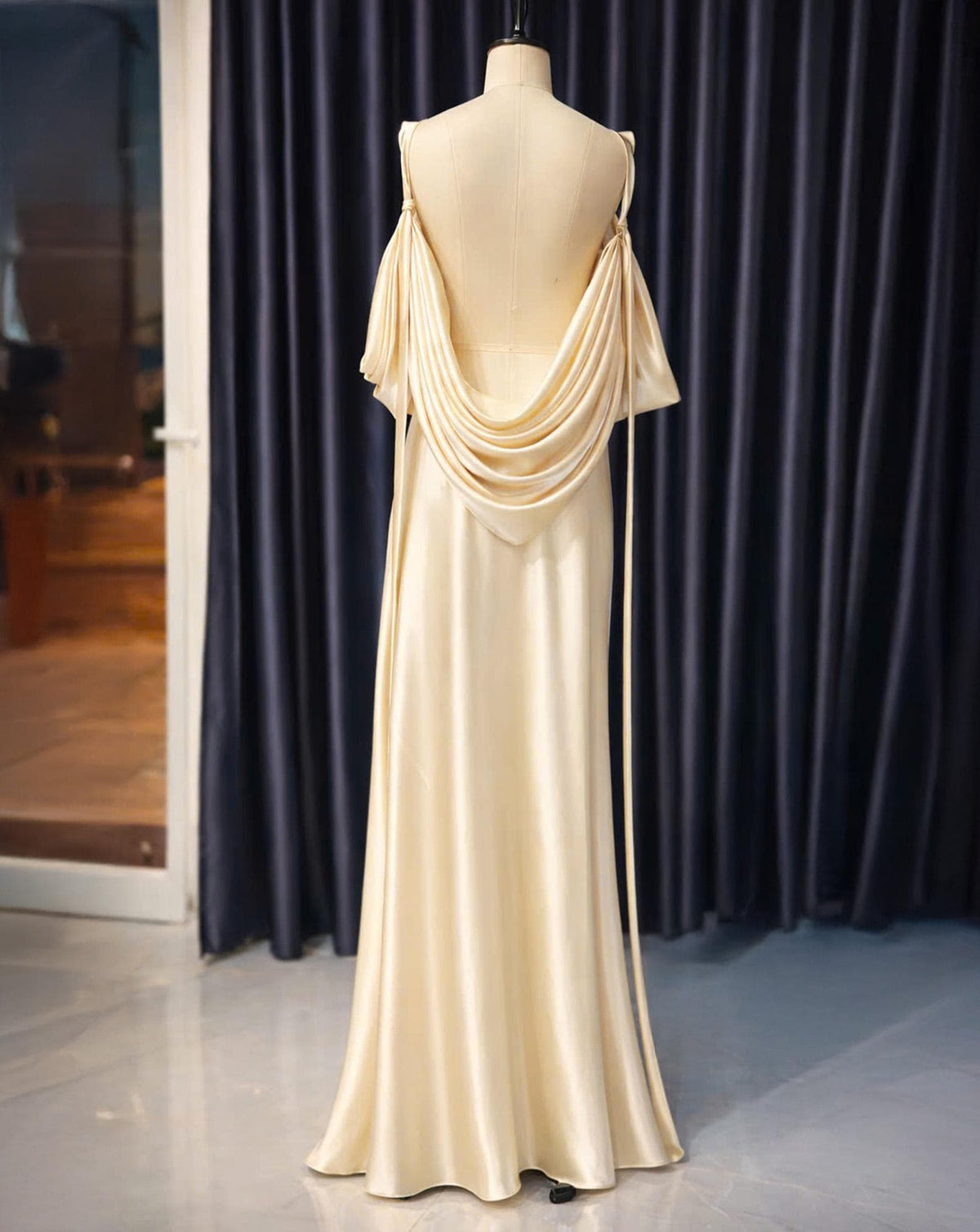May include: A cream-colored, floor-length gown on a mannequin. The dress features a draped cowl neckline and off-the-shoulder sleeves. The back is open, with delicate straps. The fabric has a subtle sheen, and the dress is displayed against a dark blue curtain.