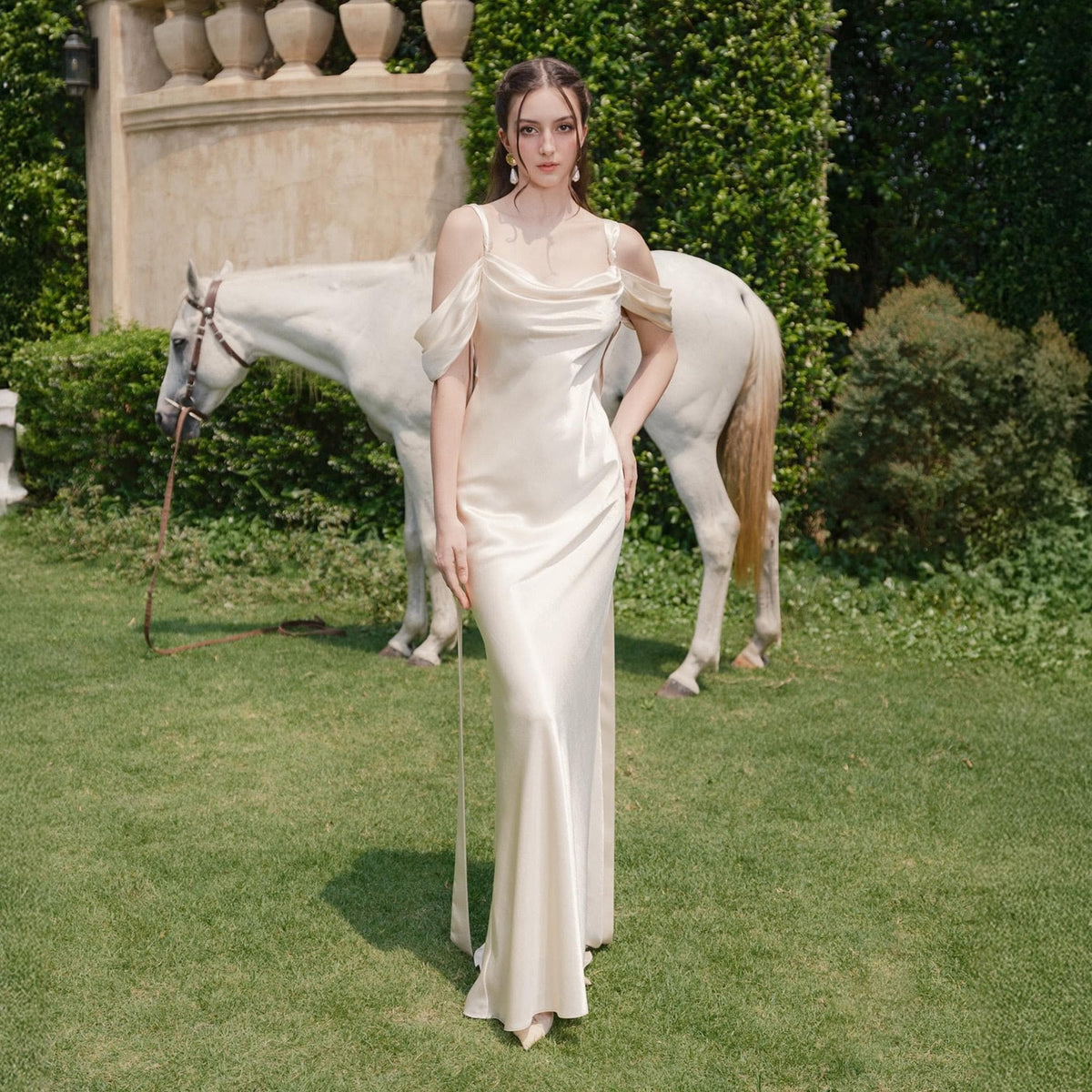 May include: A cream-colored, floor-length satin gown with a draped cowl neckline and off-the-shoulder sleeves. The dress is worn outdoors with a white horse in the background. The dress has a simple, elegant design.
