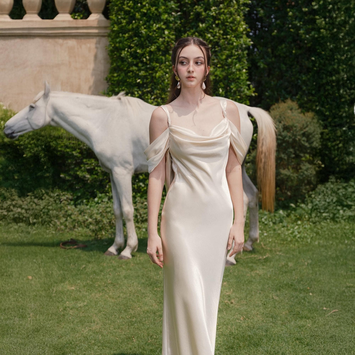 May include: A cream-colored, floor-length dress with a cowl neckline and draped sleeves. The dress is worn outdoors in front of a white horse and green foliage. The dress has a simple, elegant design.