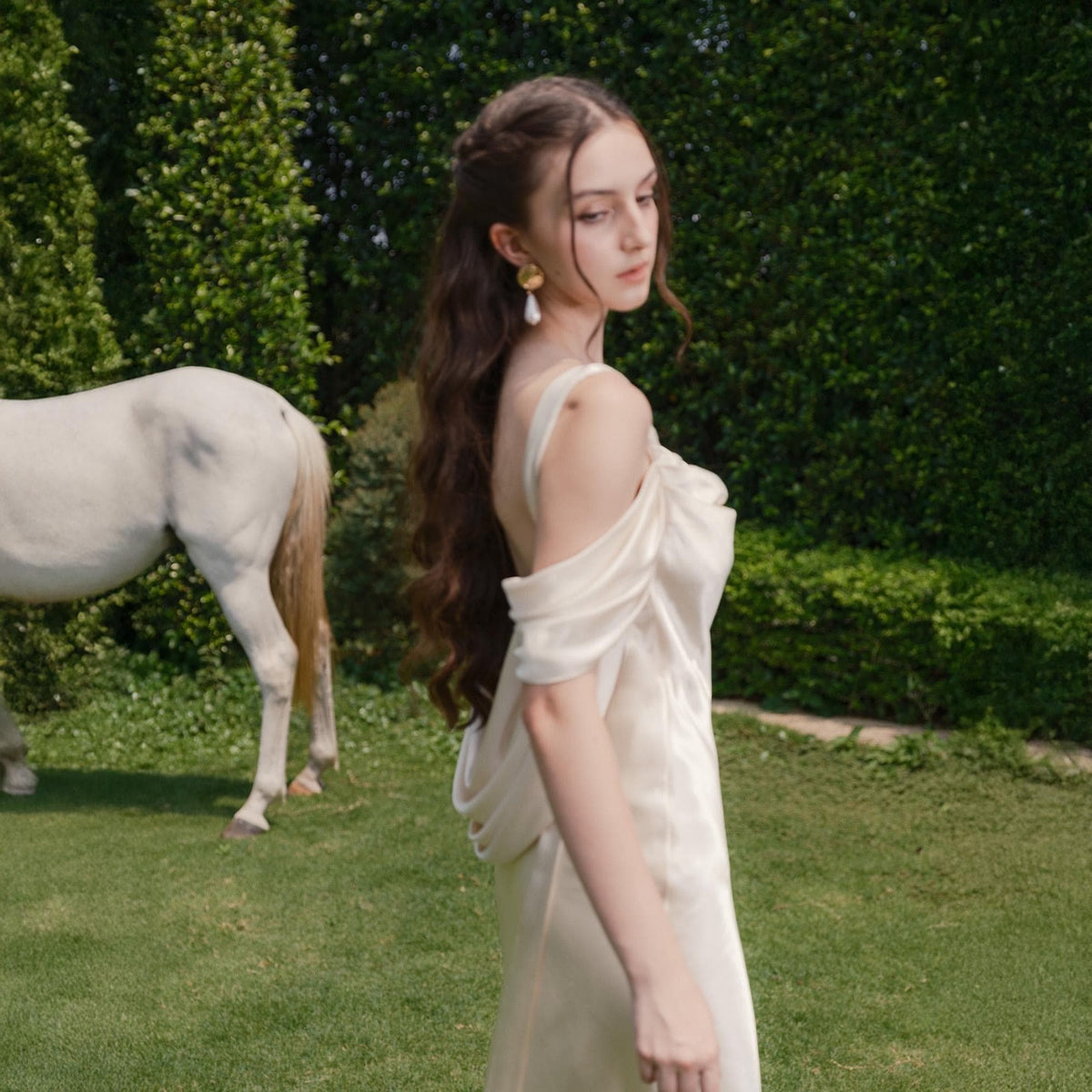 May include: A woman in an off-white, flowing gown with draped sleeves stands on a grassy lawn. Her long, wavy brown hair cascades down her back. A white horse is partially visible in the background, with a green hedge behind them.