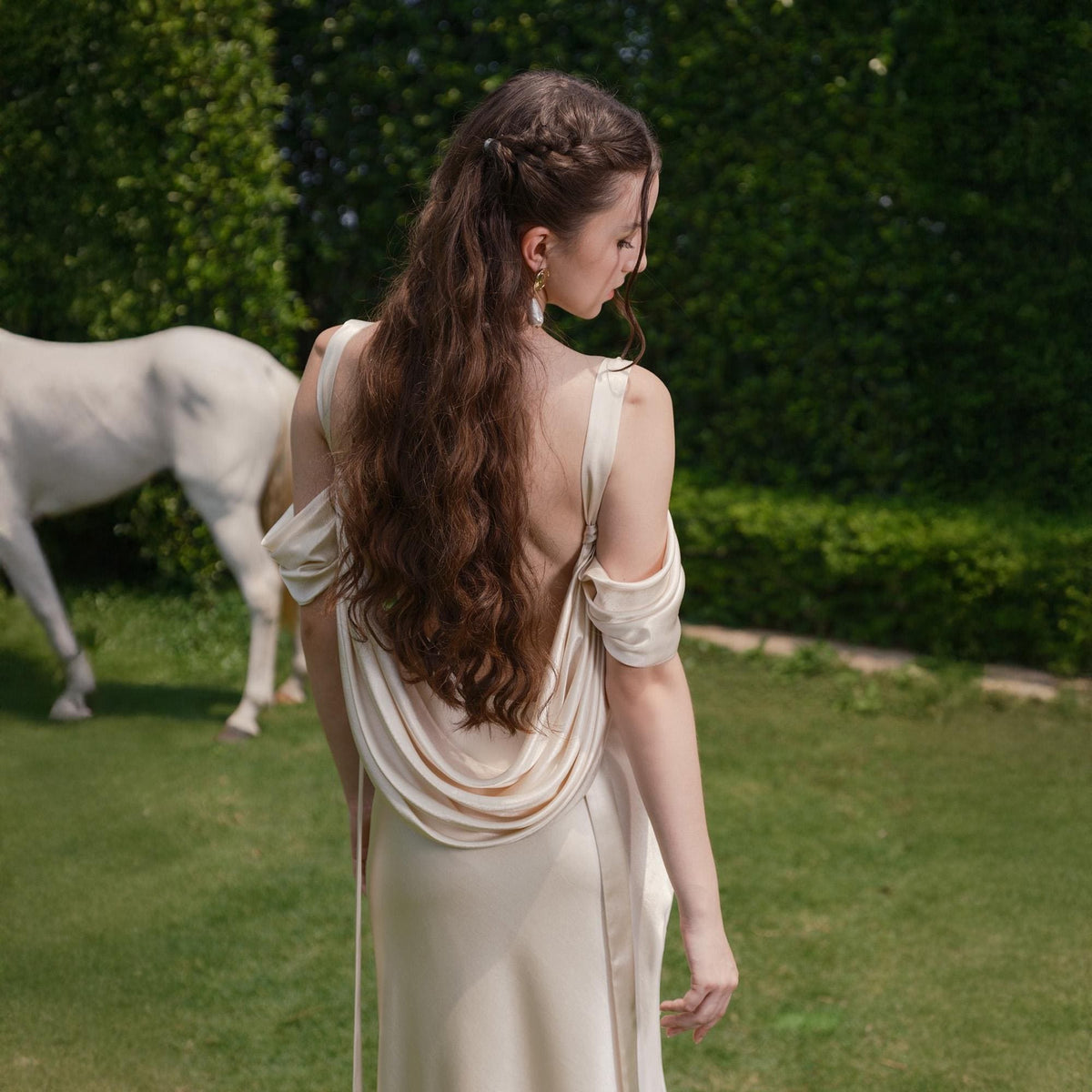 May include: A cream-colored, off-the-shoulder dress with a draped back detail. The dress has a flowing silhouette and is worn by a person with long, wavy brown hair. A white horse is in the background, standing on green grass.