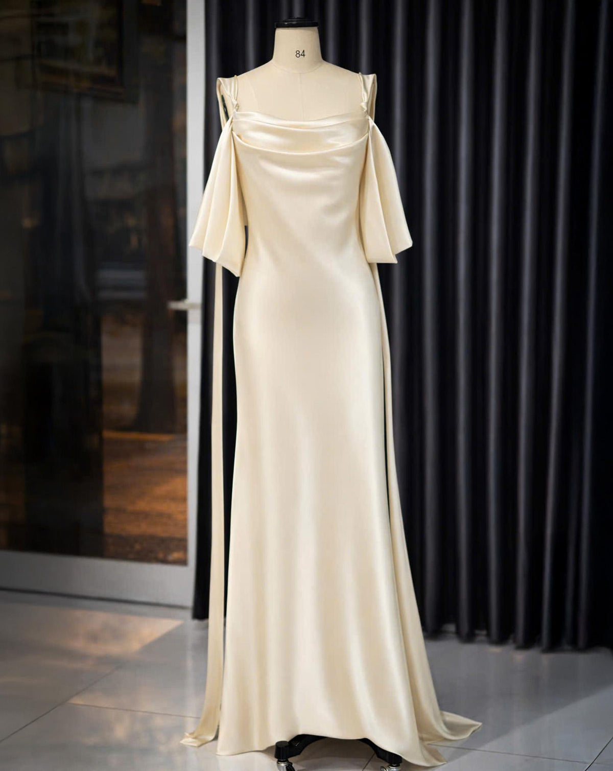 May include: A cream-colored, floor-length gown with a draped neckline and off-the-shoulder sleeves. The dress features a flowing skirt and delicate straps. The dress is displayed on a mannequin.