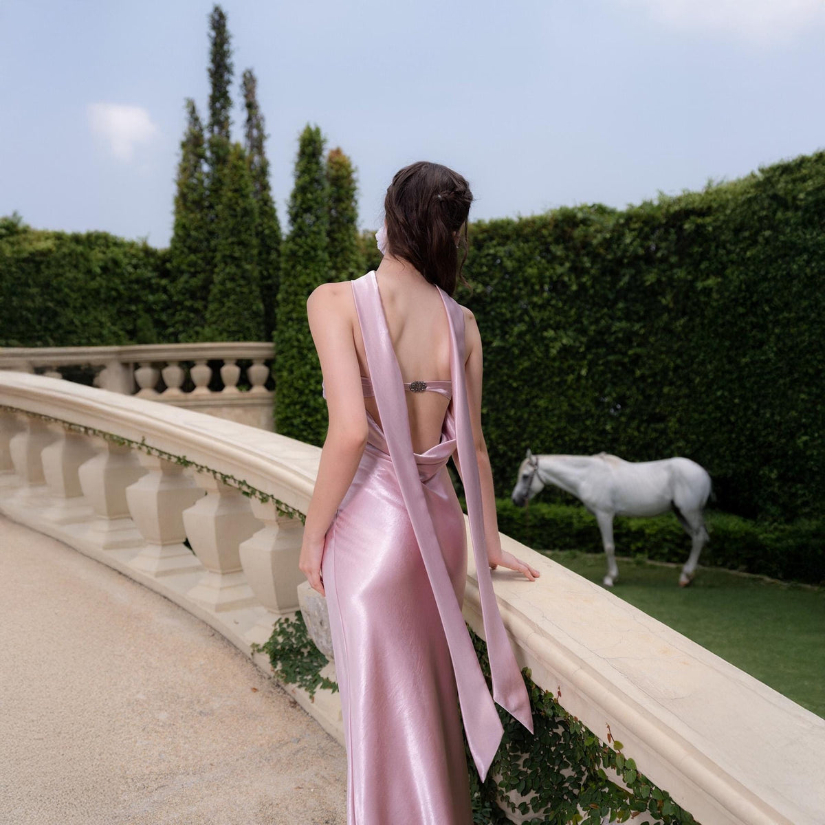 May include: A light pink satin gown with a halter neckline and open back. The dress features long, flowing fabric that drapes down the back. A white horse stands in the background near a green hedge.