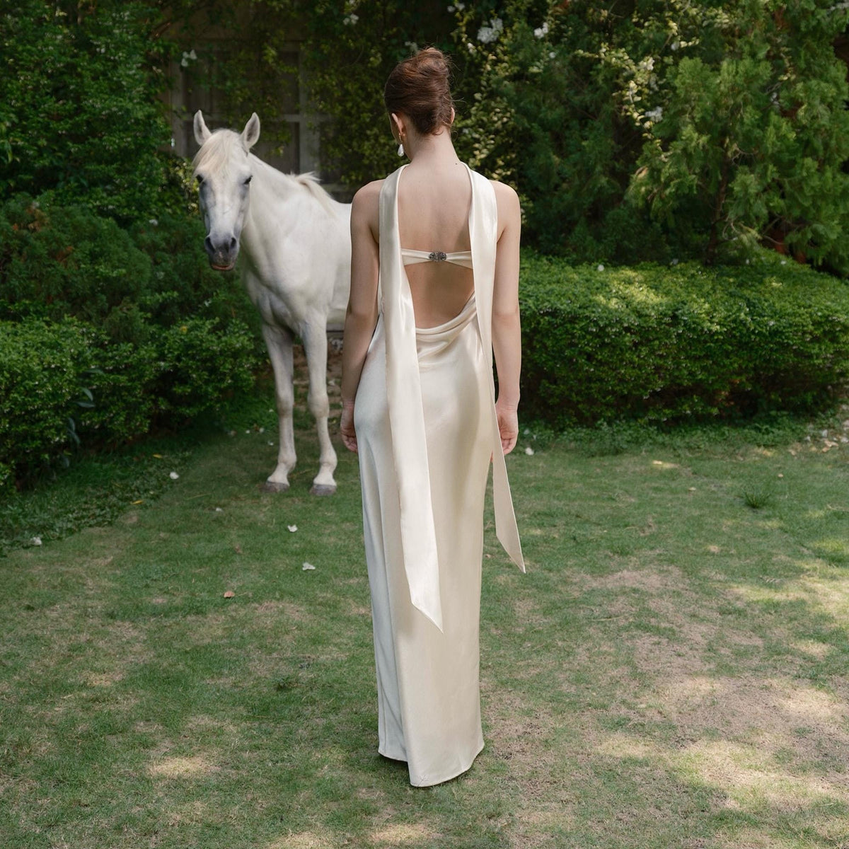 May include: A cream-colored, floor-length gown with a low back and flowing fabric. The dress features a halter-style neckline and a decorative clasp at the back. A white horse stands in the background on a grassy lawn, surrounded by green foliage.