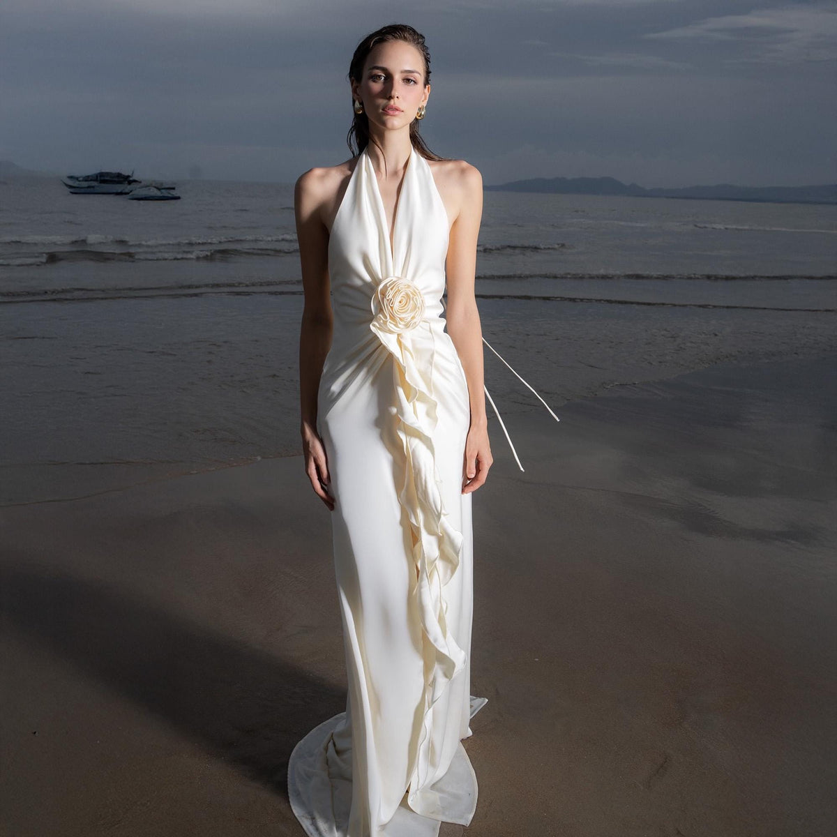 May include: A white halter-neck gown with a flowing skirt and a floral accent at the waist. The dress features a cascading ruffle detail down the front. The model is standing on a beach with the ocean and a cloudy sky in the background.
