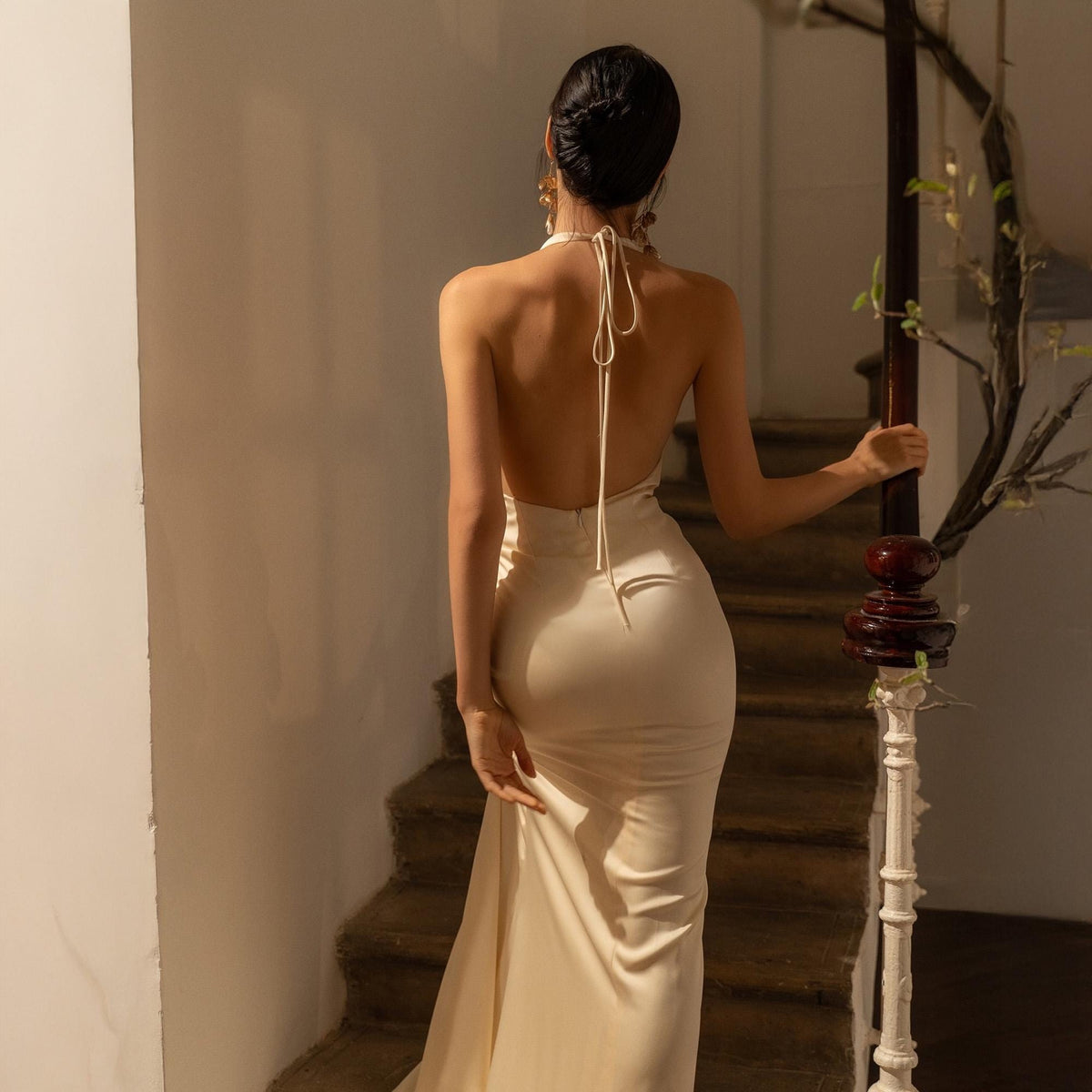 May include: A cream-colored, backless halter dress with a flowing skirt. The dress is worn by a person ascending a staircase with a wooden banister. The dress has a subtle sheen, suggesting a satin or silk fabric.