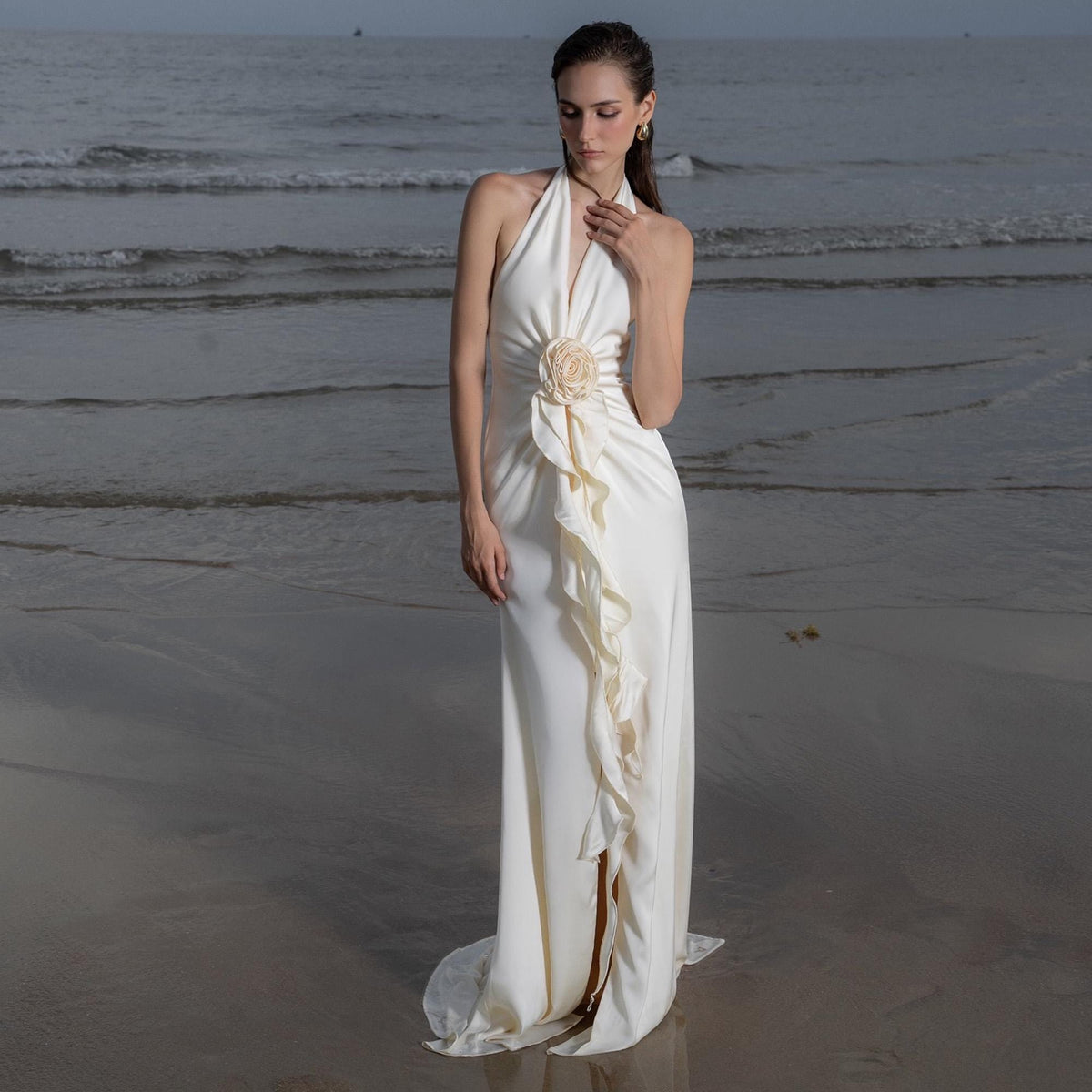 May include: A white halter-neck gown with a floral embellishment at the waist and a flowing ruffle detail. The dress has a high slit and a train, perfect for a beach wedding or formal event. The model is standing on a sandy beach with the ocean in the background.