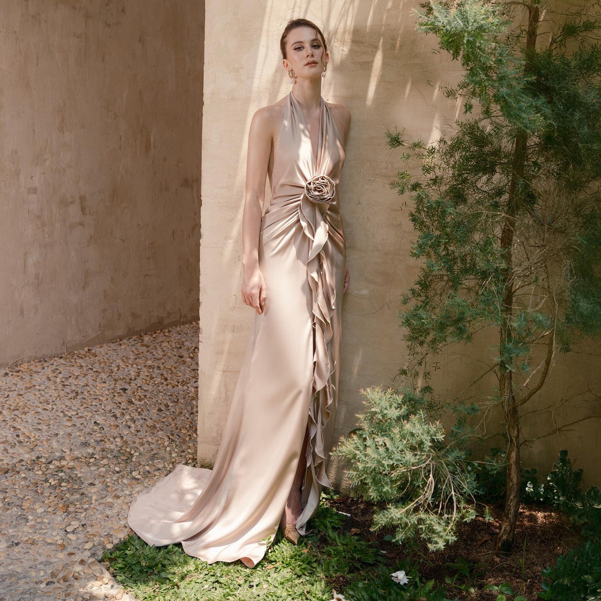 May include: A floor-length, champagne-colored gown with a halter neckline and a flowing skirt. The dress features a floral embellishment at the waist and cascading ruffles down the side. The dress has a train and is made of a silky material.