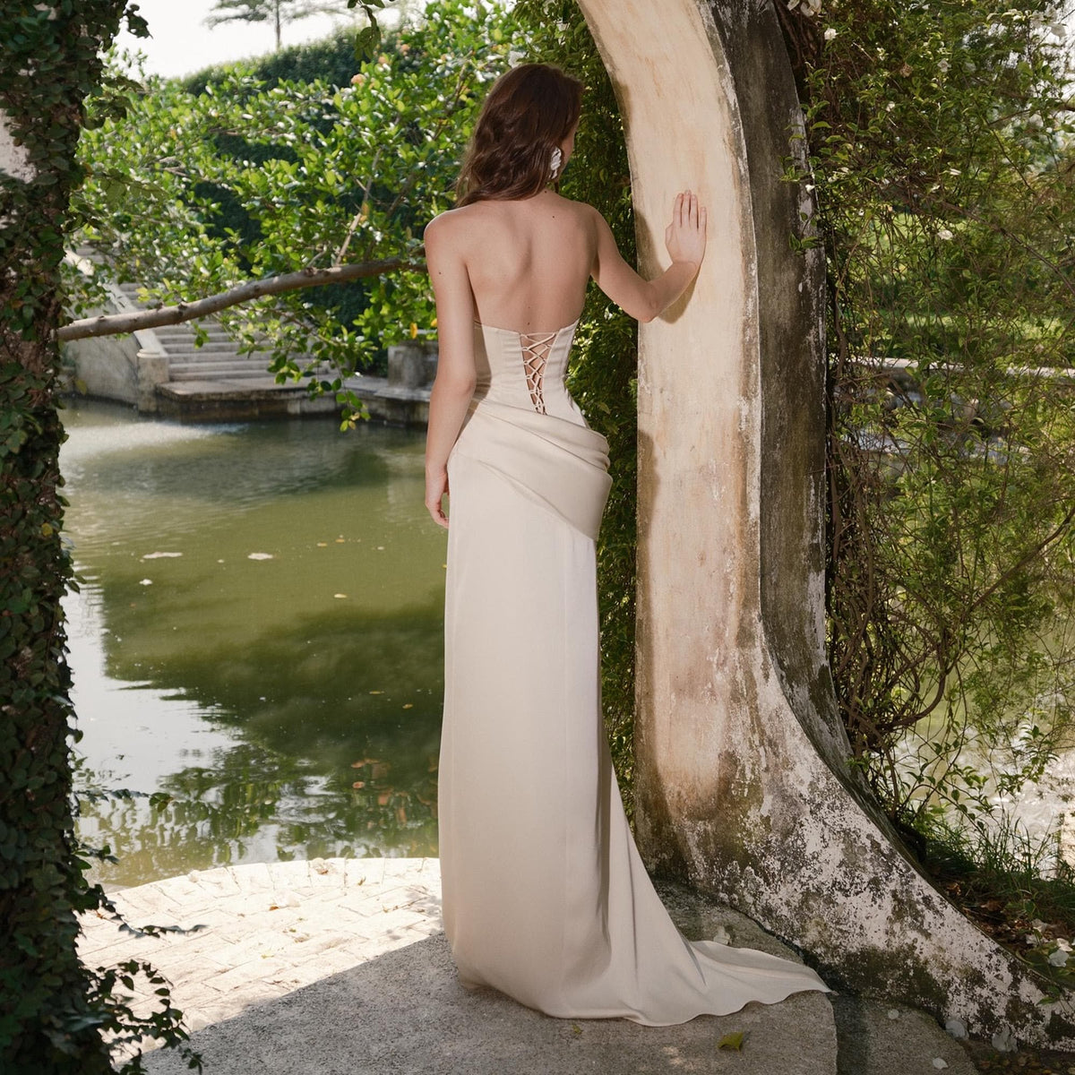 May include: A strapless, cream-colored gown with a corset back and a flowing skirt. The dress features a draped design at the waist and a subtle train. The woman is standing near a body of water, with greenery in the background.